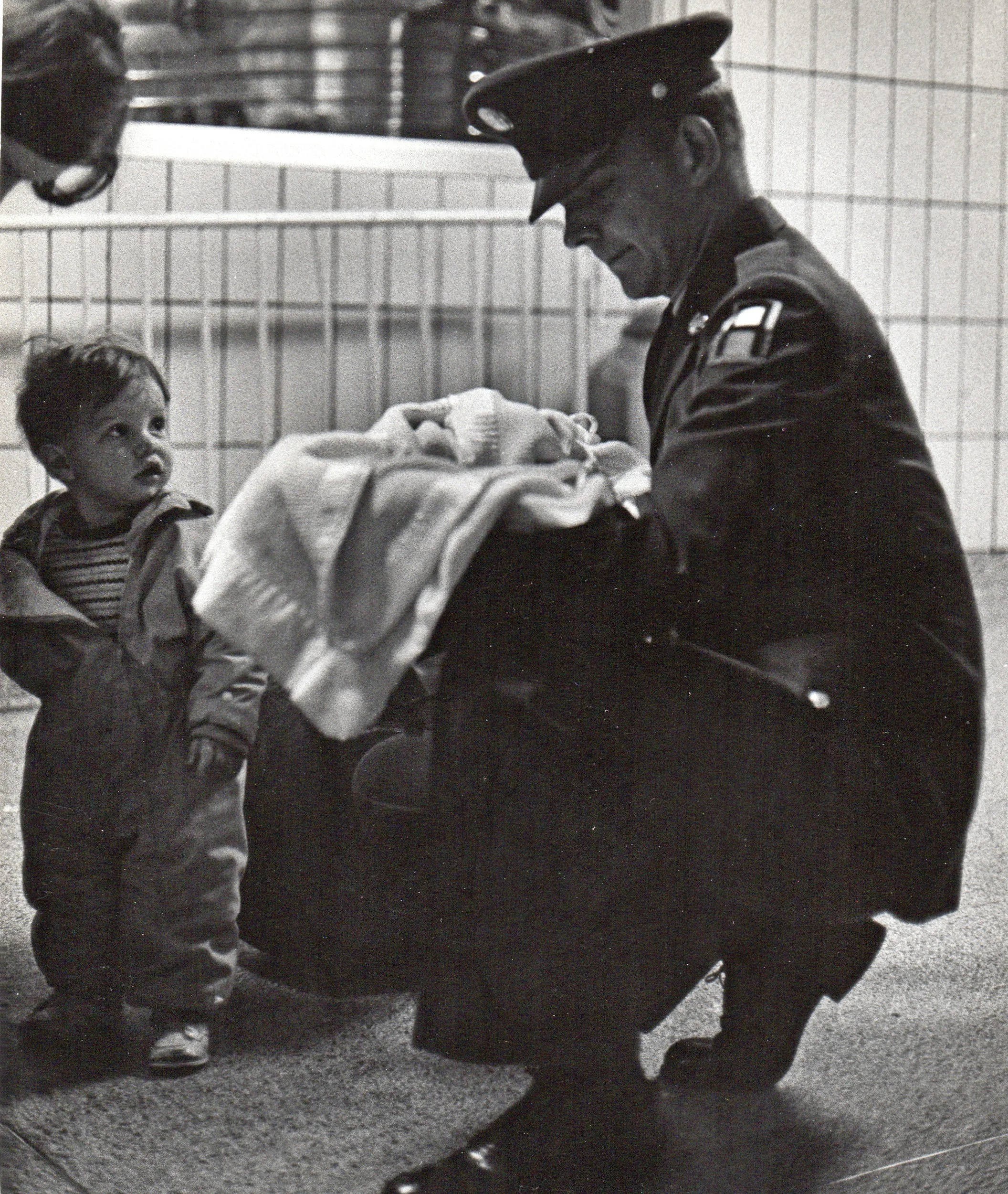 A police officer dressed in uniform kneeling down and holding a newborn wrapped in a blanket, with a young child looking on curiously at the scene, in an indoor setting.