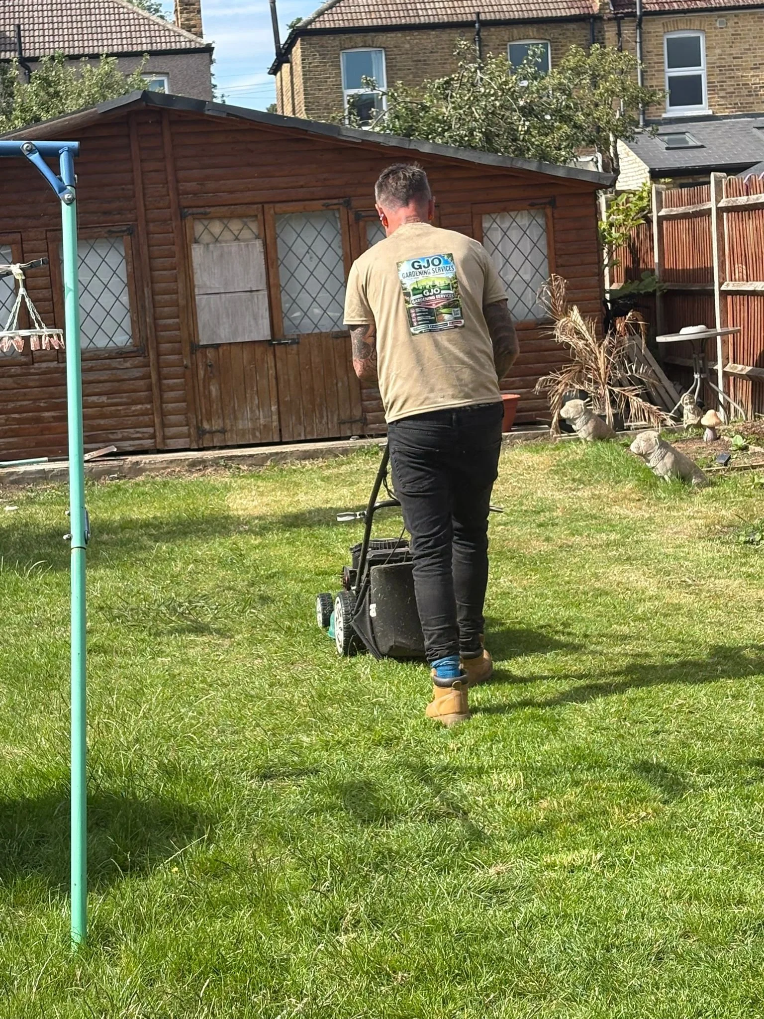 Man with short hair and tattoos wearing a tan GJO Gardening Services t-shirt, black pants, and tan boots, working with a lawnmower in a backyard with a wooden shed and a wooden fence.