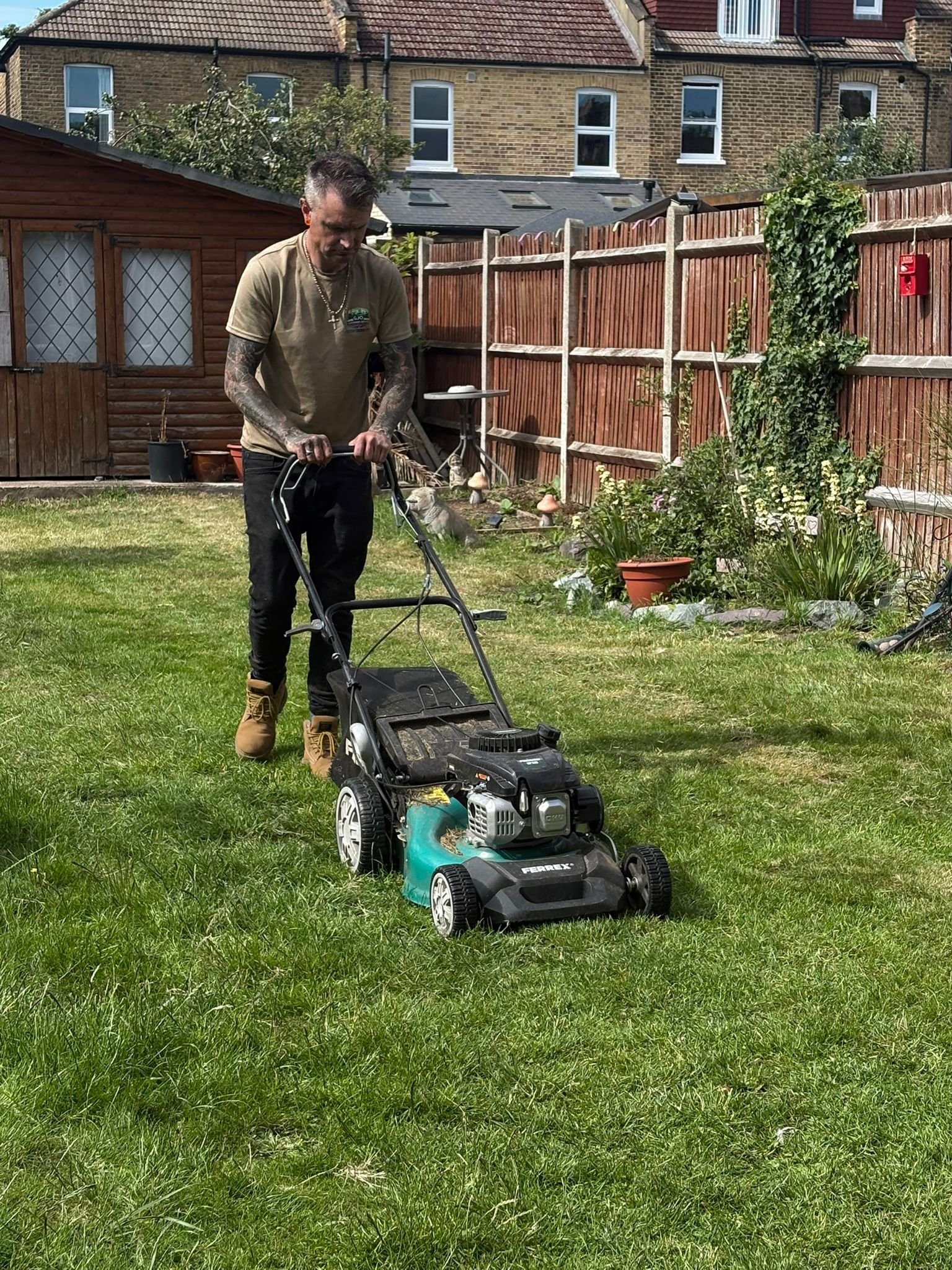 A man with tattoos on his arms wearing a beige T-shirt and tan work boots is pushing a lawnmower on a grassy backyard. There are potted plants, a wooden fence, and a small wooden shed in the background, with residential brick houses visible behind the fence.