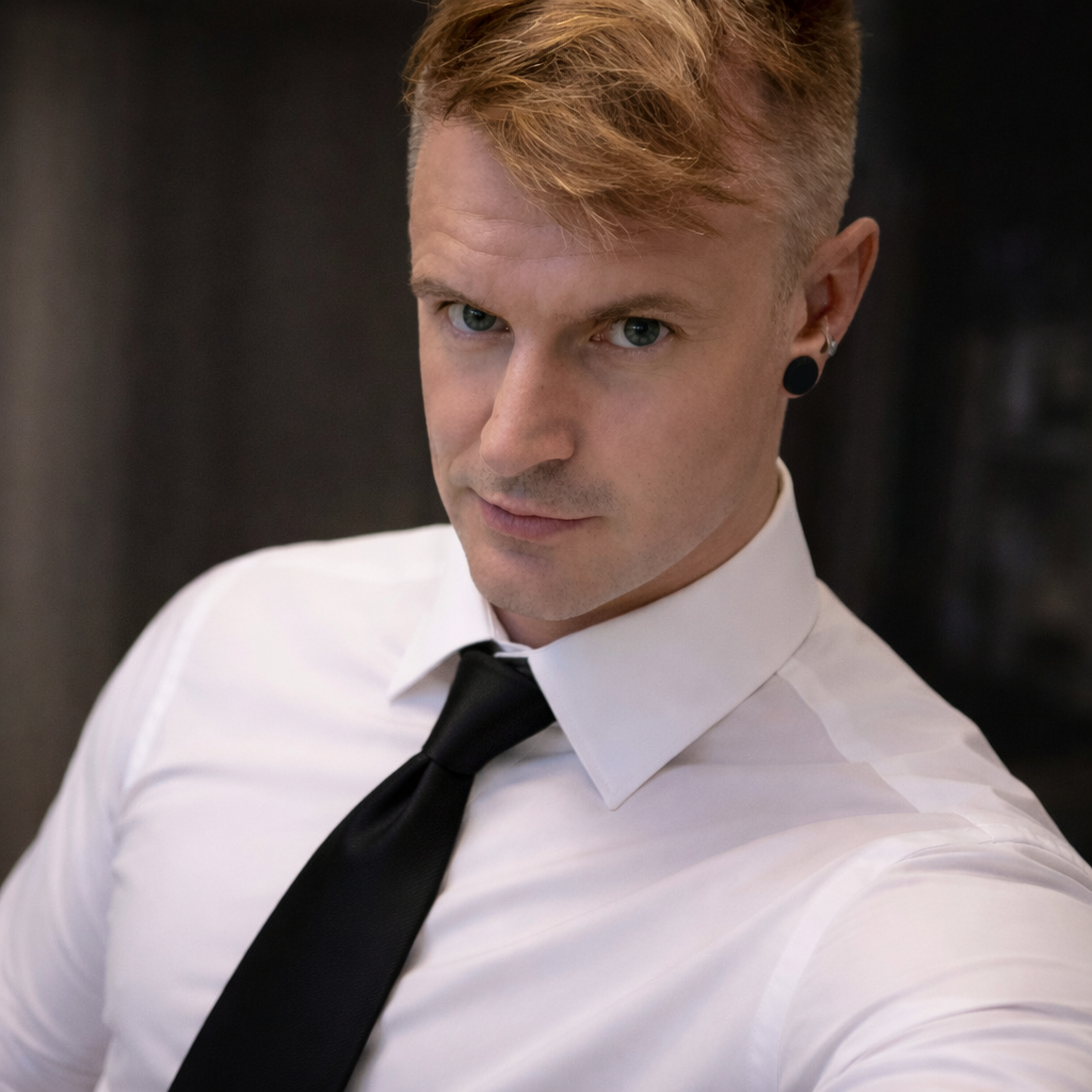 A young man with blond hair and blue eyes wearing a white dress shirt, black tie, and black ear gauge earrings, looking into the camera with a neutral expression.