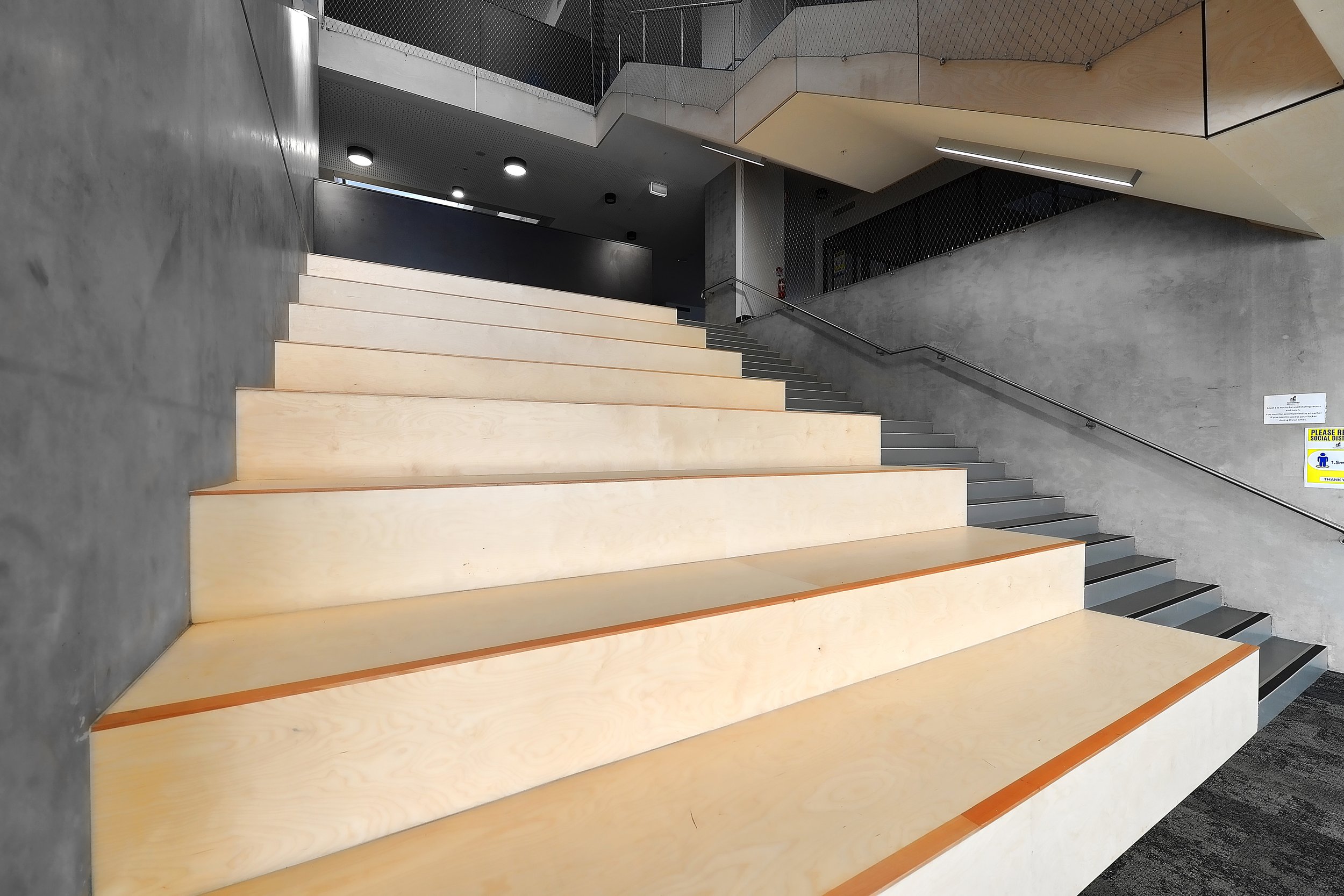Interior view of a modern building with wide wooden stairs, concrete walls, and metal railings.