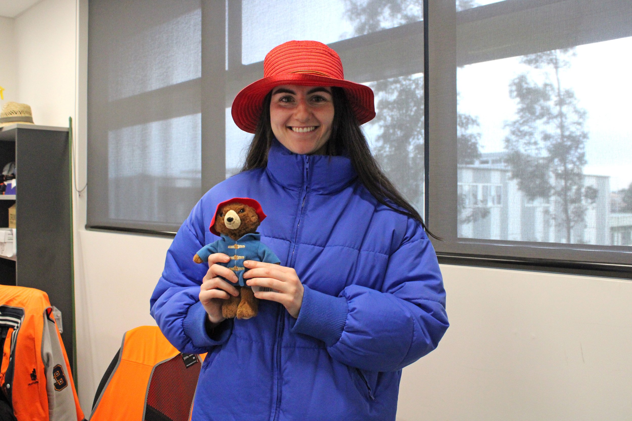 A woman in a red hat and a blue jacket smiling and holding a teddy bear dressed in a blue coat.