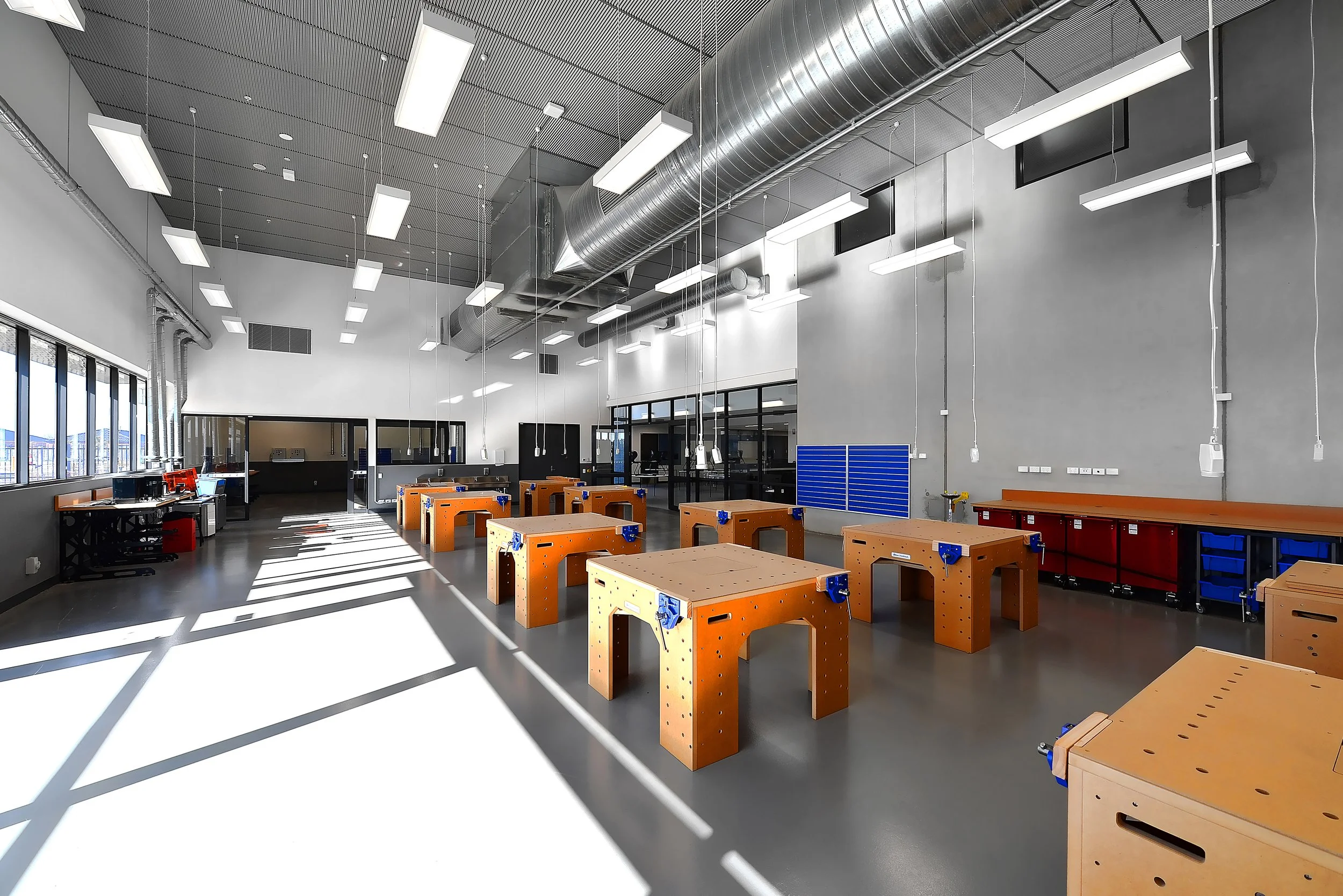 A modern workshop or makerspace with orange workbenches arranged in a grid. The space has large windows, gray walls, and a high ceiling with exposed ductwork and lighting fixtures. There are tools and equipment along the walls.