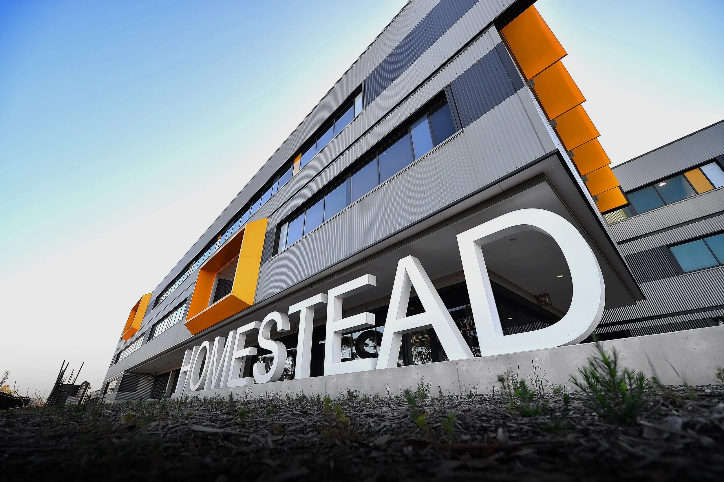 Modern multi-story building with large sign reading 'HOMESTEAD' in front, featuring gray siding, orange accents, and large windows.