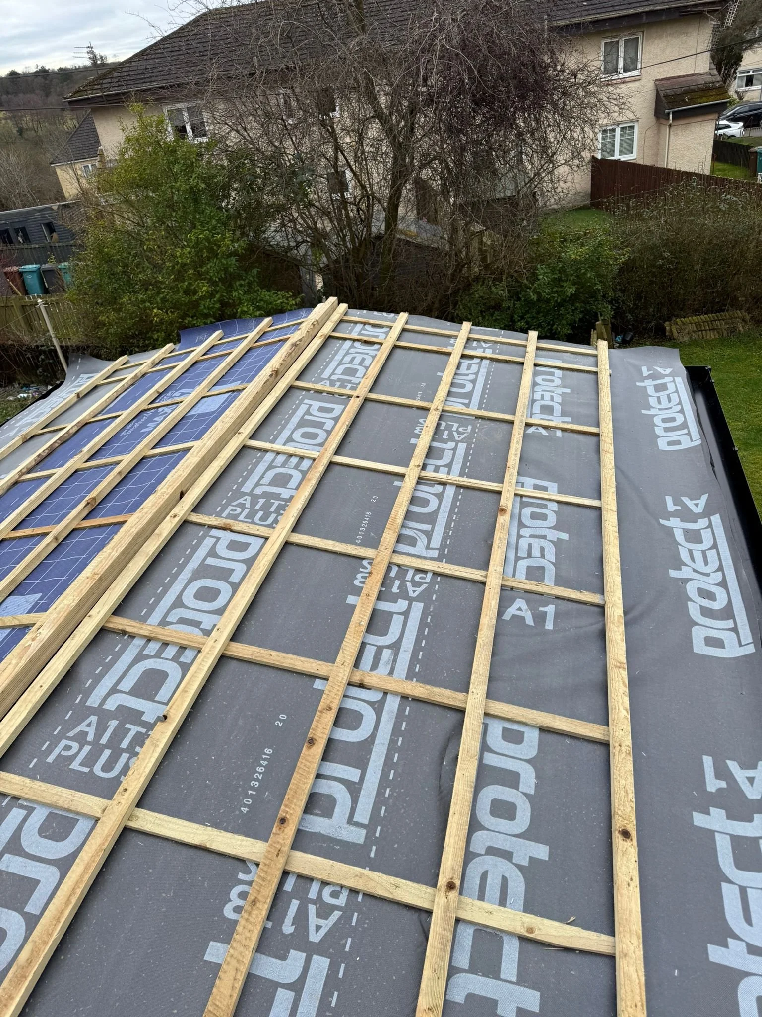Construction site on a roof with wooden framing and underlayment material, overlooking a suburban neighborhood with trees and houses.