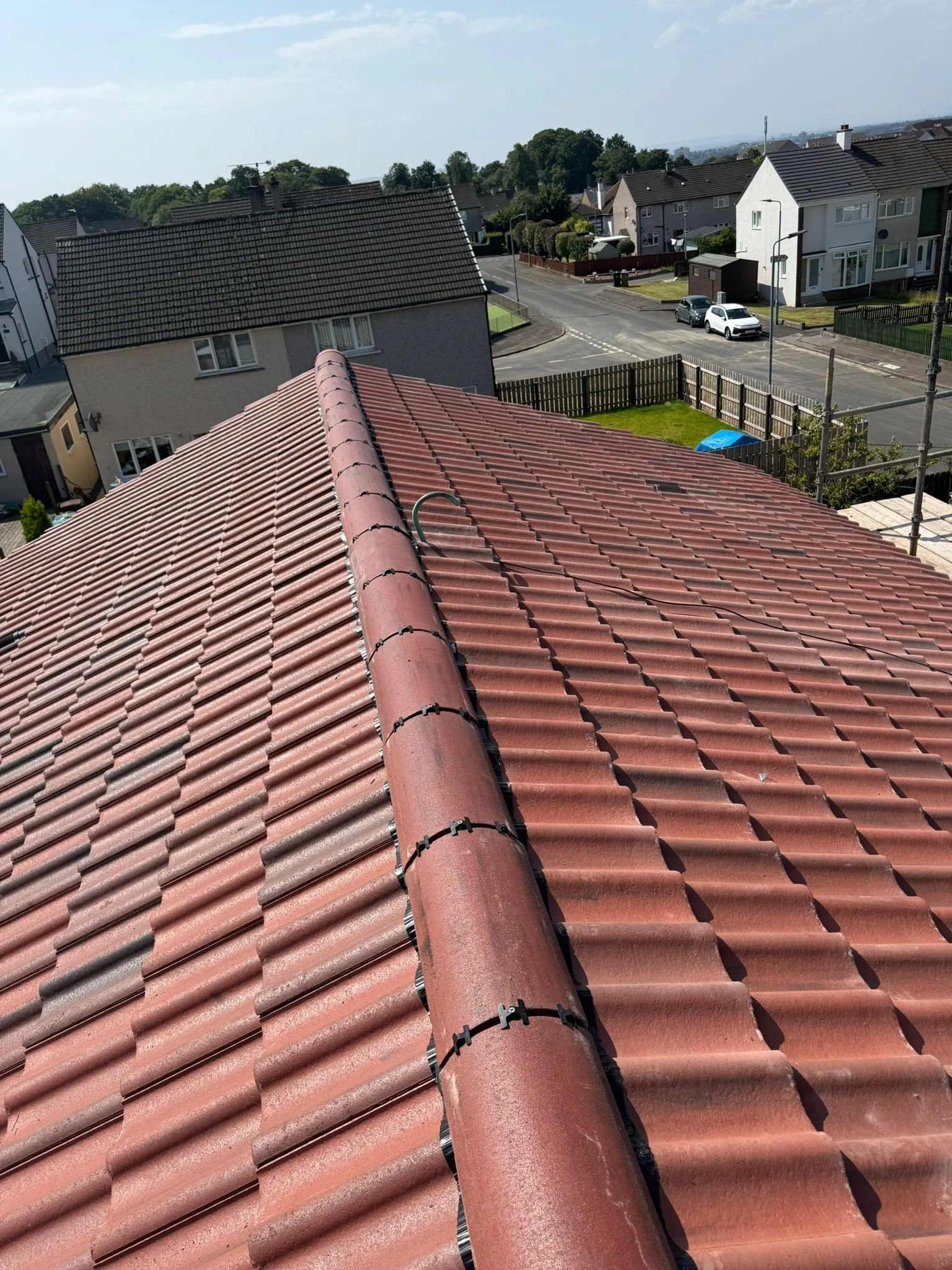 View from the roof of a residential house showing red tiles and a ridge vent, with neighboring houses, street, and parked cars in the background.