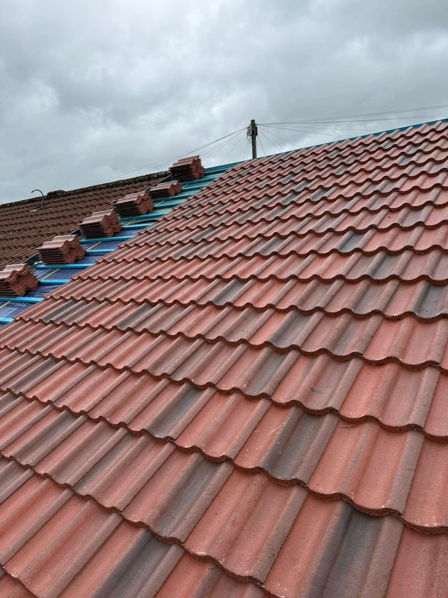 Red clay roof tiles on a house with some tiles being replaced or repaired, with a cloudy sky and overhead power lines visible in the background.