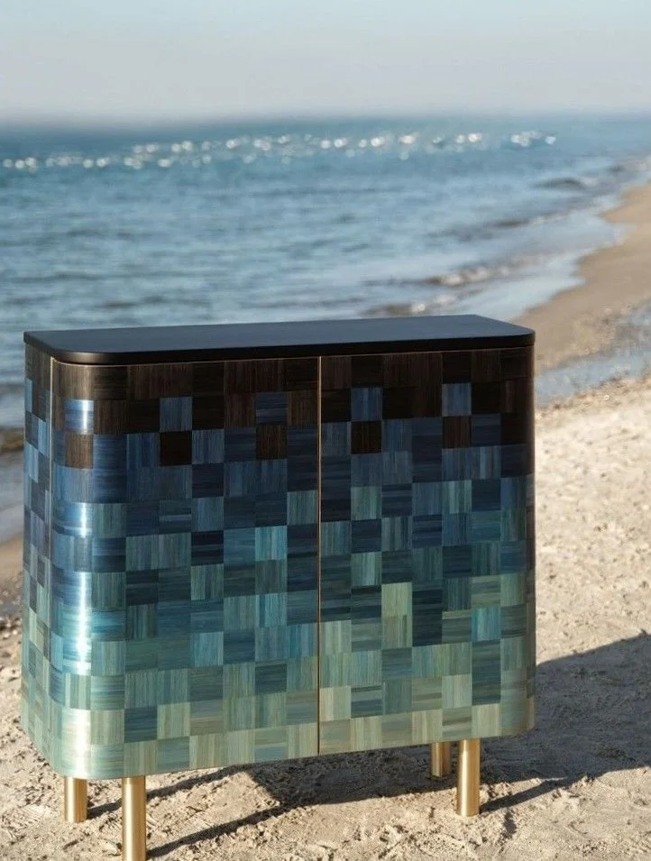 Decorative cabinet with a colorful checkered pattern stands on the sandy beach near the ocean, under clear sky.