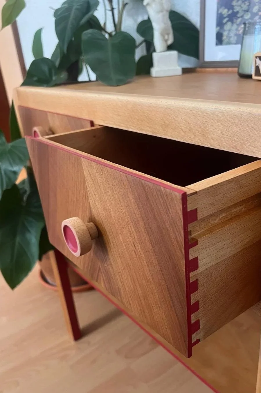 A wooden drawer partially pulled open on a wooden dresser with decorative items and a framed picture on top, and green houseplants nearby.