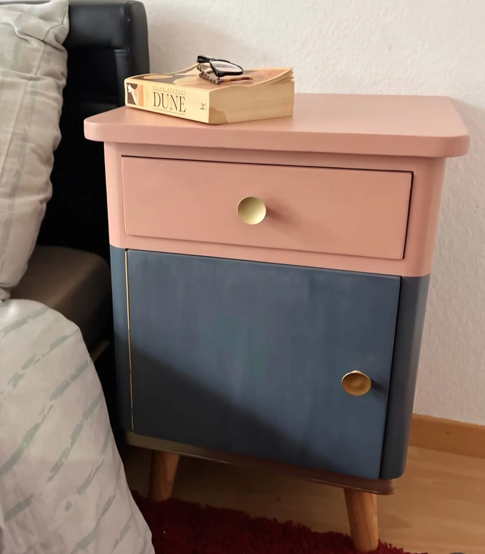 A colorful modern nightstand with two drawers, pink on top and blue below, with round gold-colored handles, next to a bed with a striped bedspread and a beige pillow.