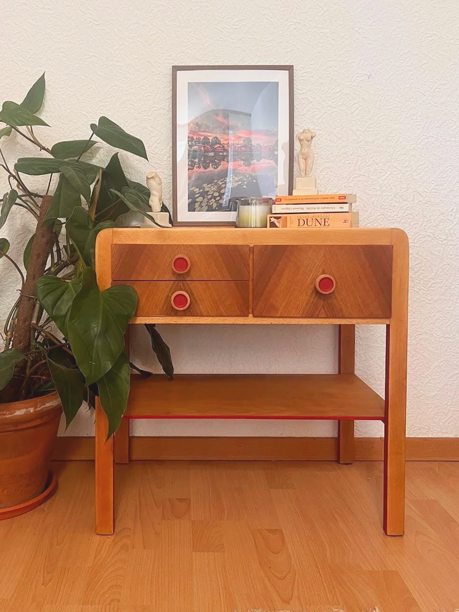 A wooden console table with four drawers, four red knobs, and an open shelf at the bottom, placed against a textured white wall. On top of the table, there are books, a small sculpture, a candle, and a framed landscape photograph. To the left, there is a large potted plant with green leaves.