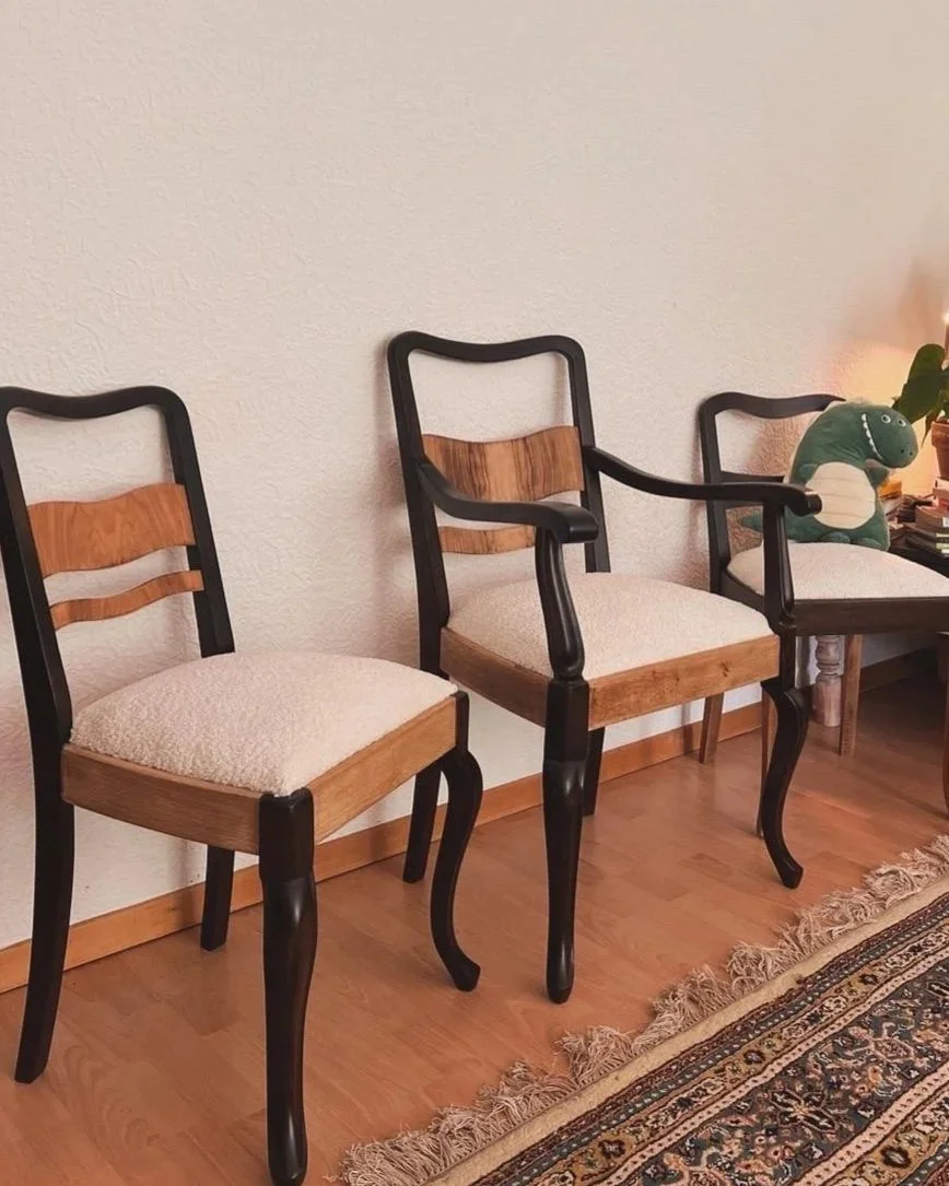 Three chairs with wooden seats and backs, black frames, and curved legs, placed against a textured white wall on a hardwood floor, with a small table to the right holding a plush dinosaur toy and a lamp.