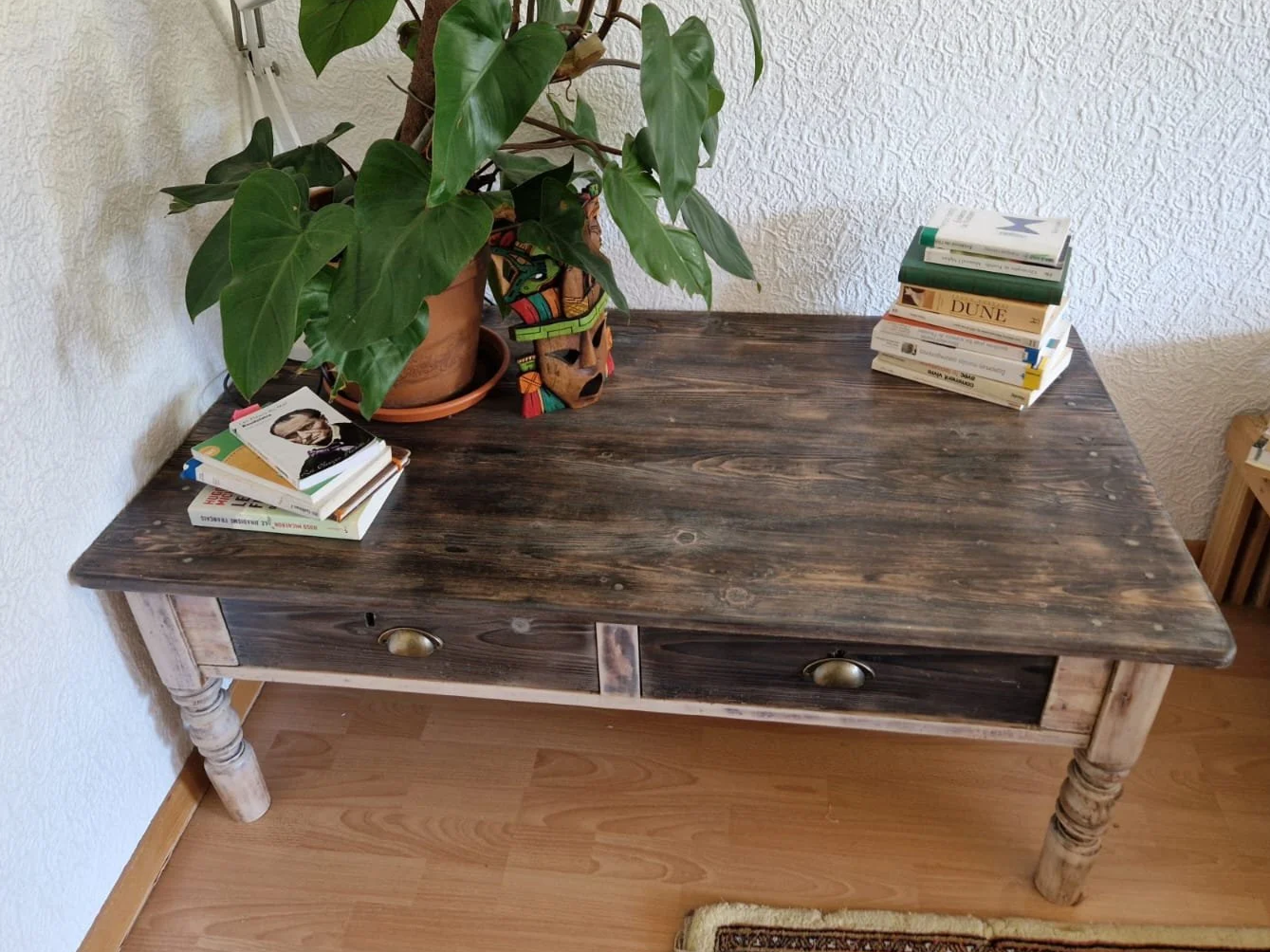 A rustic wooden table with two small drawers, topped with a potted plant, a colorful carved mask, and stacks of books, placed against a textured white wall.
