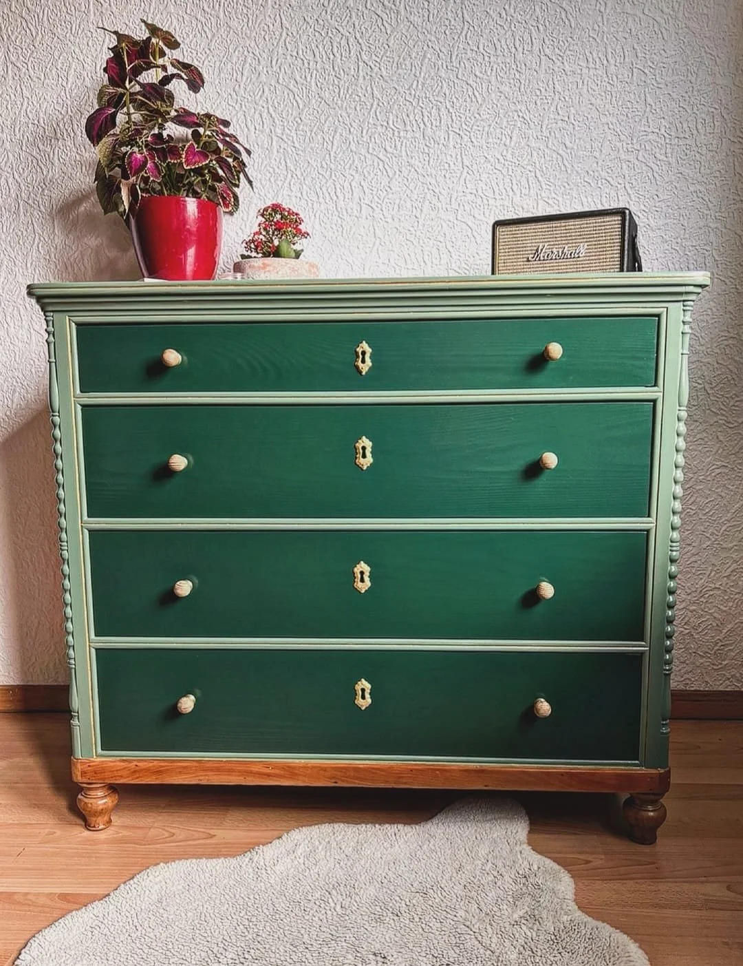A vintage green wooden dresser with five drawers and white trim, standing on a wood floor with a beige rug in front. The dresser has four wooden knobs on each drawer, a small keyhole in the center of each, a large potted plant with red and green leaves in a red pot, a small potted plant, a Marshall speaker, and another smaller item on top.