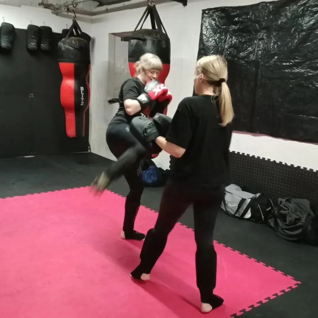Two women training in a boxing gym, one wearing boxing gloves and a focus mitt, the other in black clothes, practicing kickboxing on a pink mat.