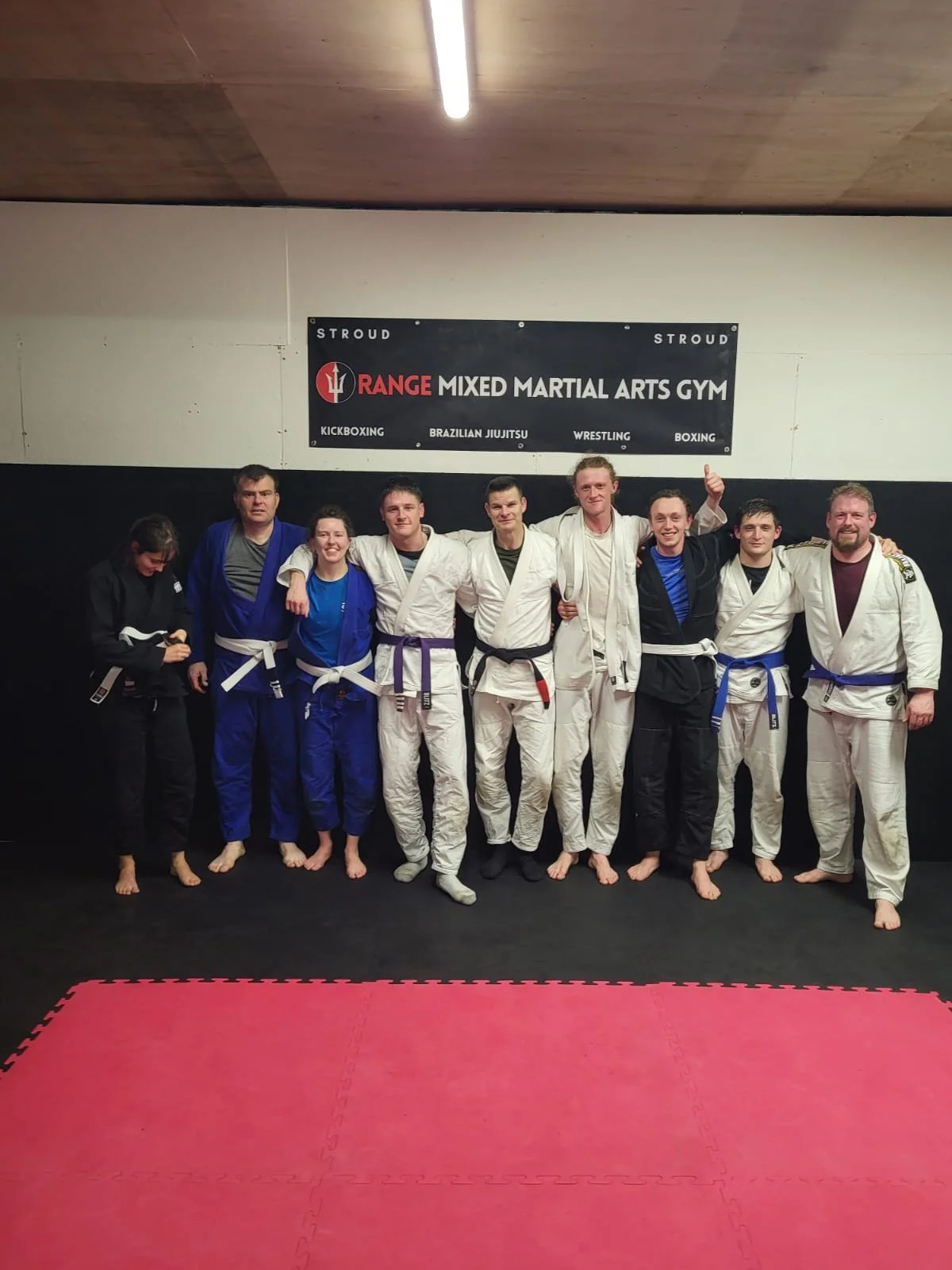 Group of martial arts students and instructors in GI uniforms standing together inside a mixed martial arts gym.