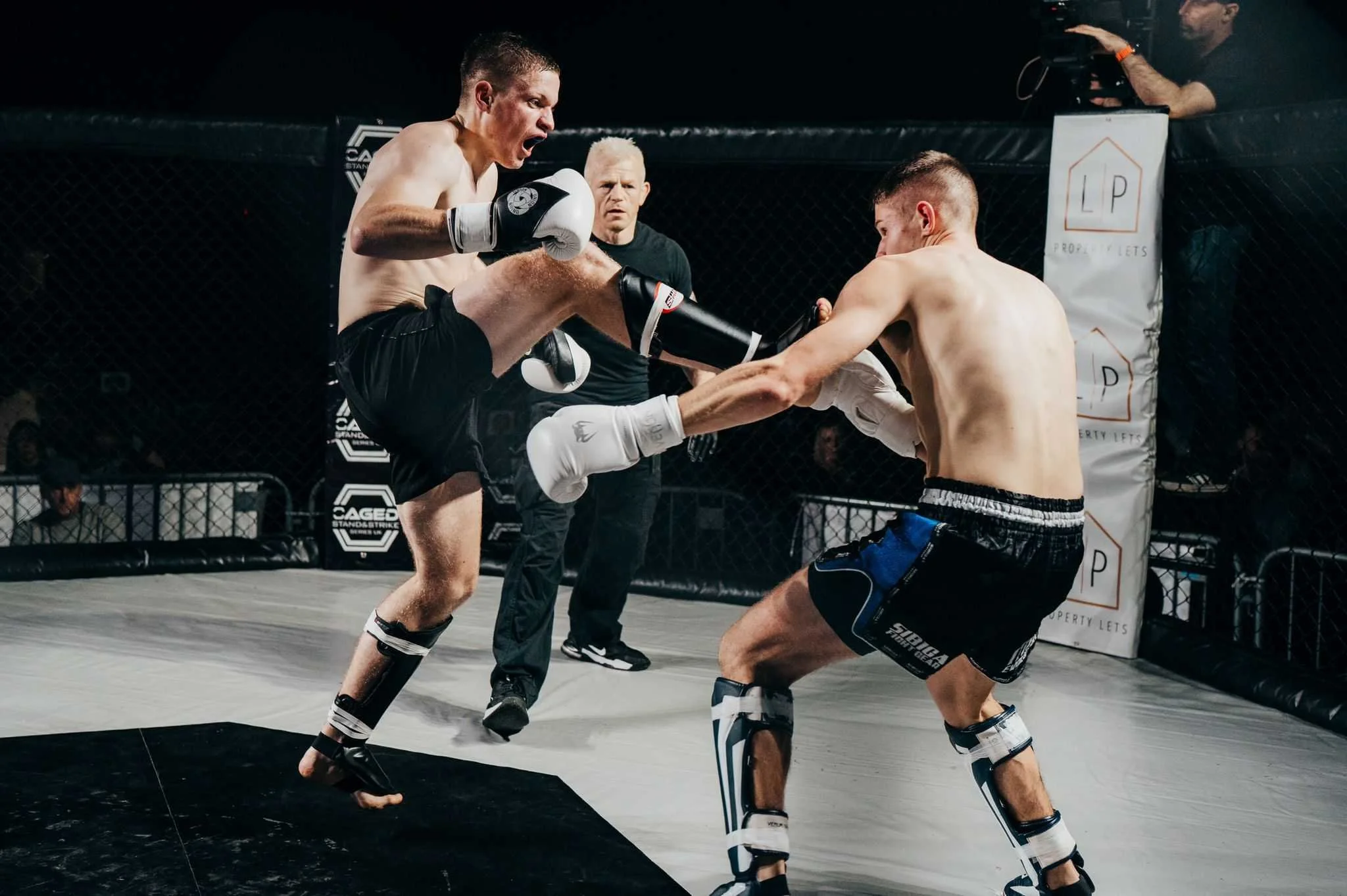 Two mixed martial artists competing in a cage fight, one delivering a kick to the other's head. A referee and cameraman are observing inside the fighting ring.