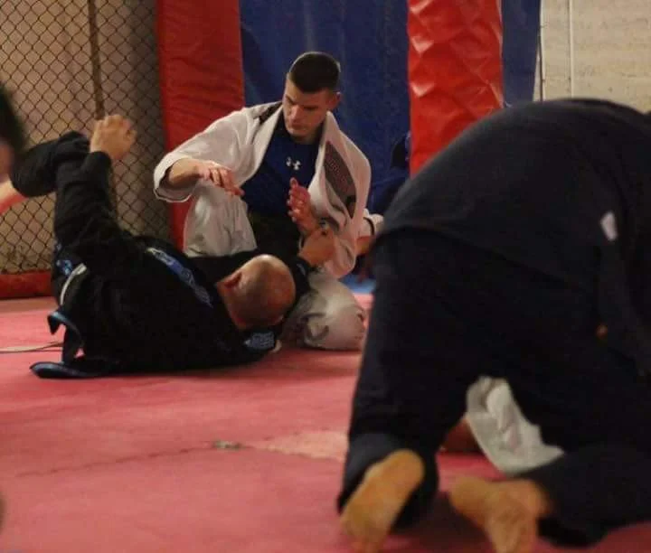Two men practicing Brazilian Jiu-Jitsu on a padded mat inside a training facility with red and blue color walls and black netting. One man in a white gi is on top, while the other in a black gi is on the ground. A third person, partially visible, is kneeling nearby.