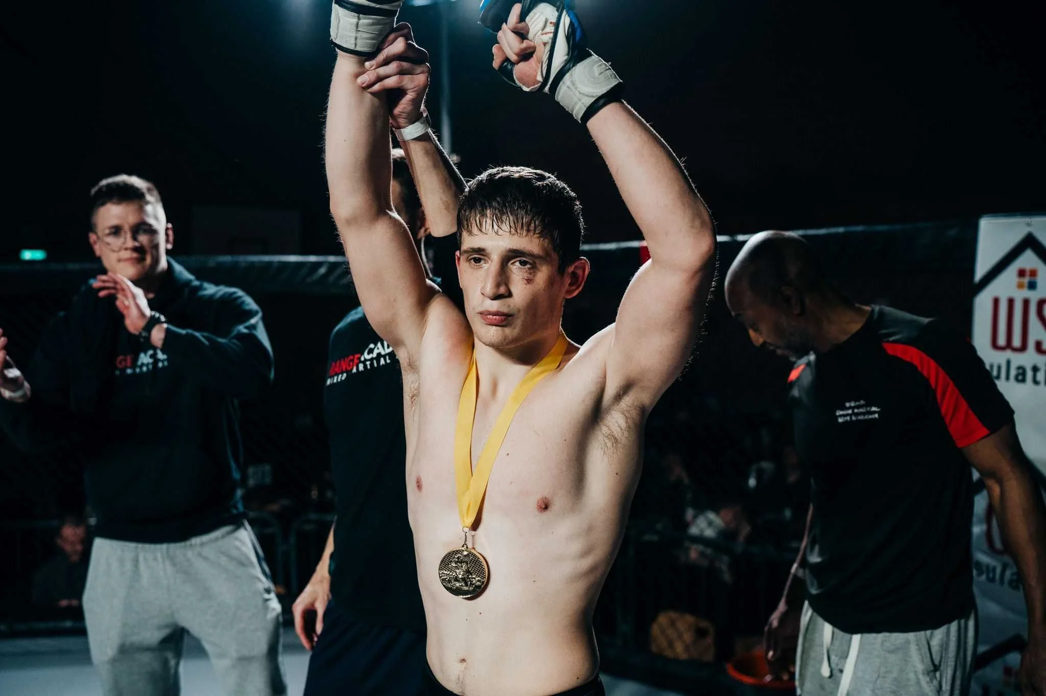 Mixed martial arts fighter standing in the octagon, holding his hands above his head, wearing a medal around his neck, with a focused expression, and surrounded by people including a referee and others in athletic gear.