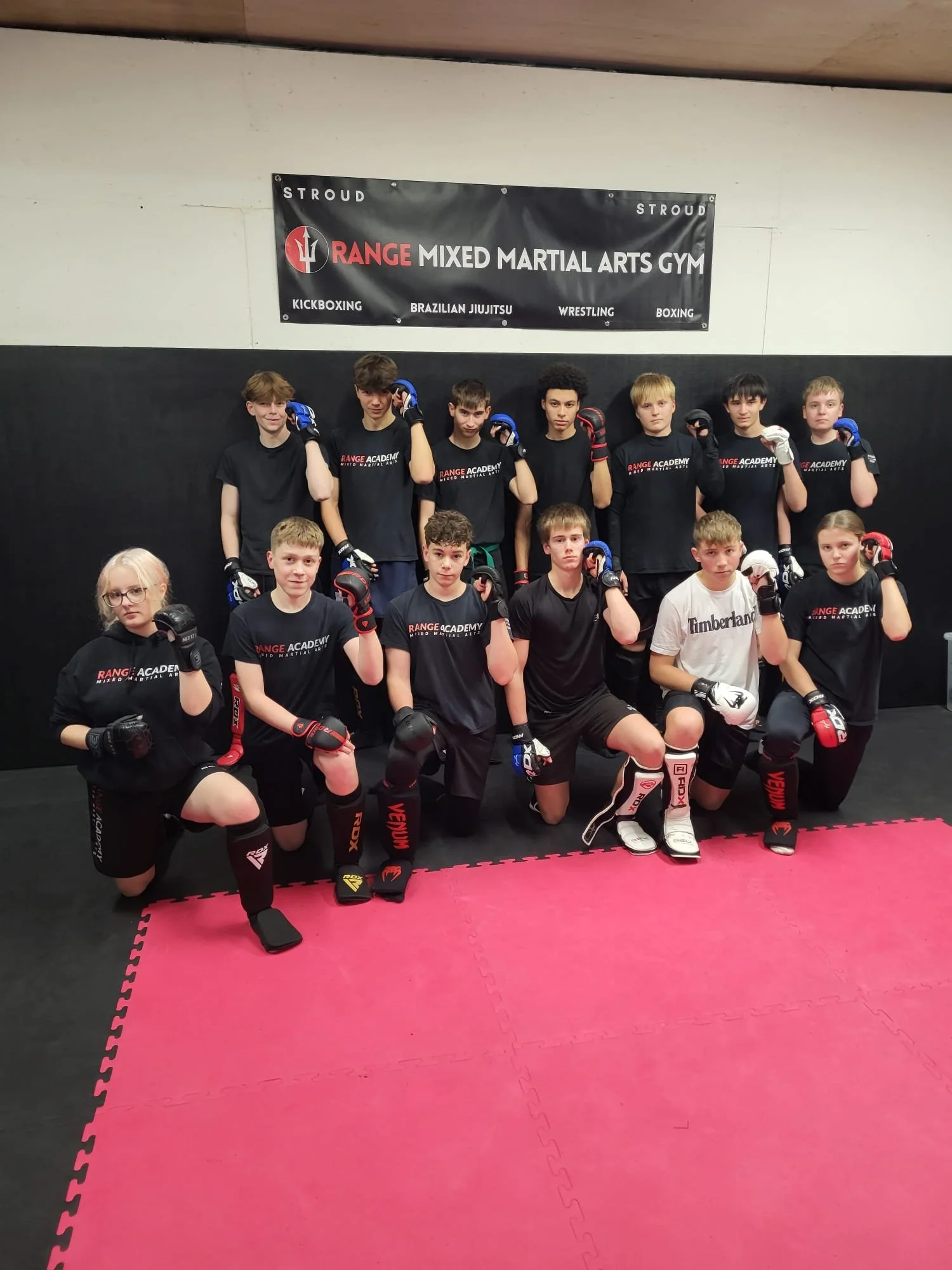 A group of young martial artists with protective gear posing for a photo in a martial arts gym. They are wearing black uniforms and are kneeling or standing on a pink and black padded floor. A banner behind them reads "Range Mixed Martial Arts Gym," indicating the martial arts training center.