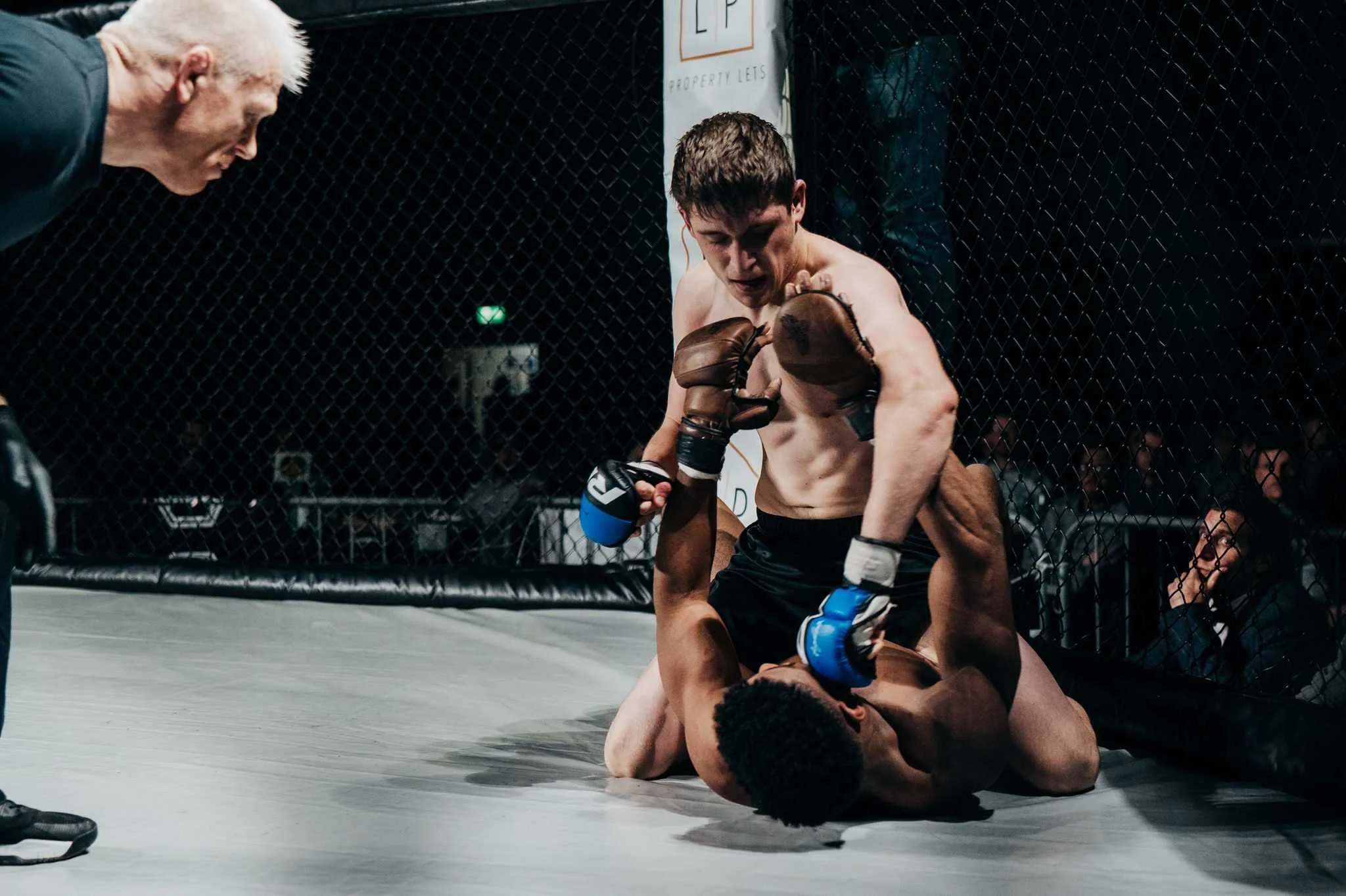 Two mixed martial arts fighters engaged in a grappling match inside a caged octagon, one fighter on top and the other on the ground, with a referee observing nearby and spectators watching from behind the cage.