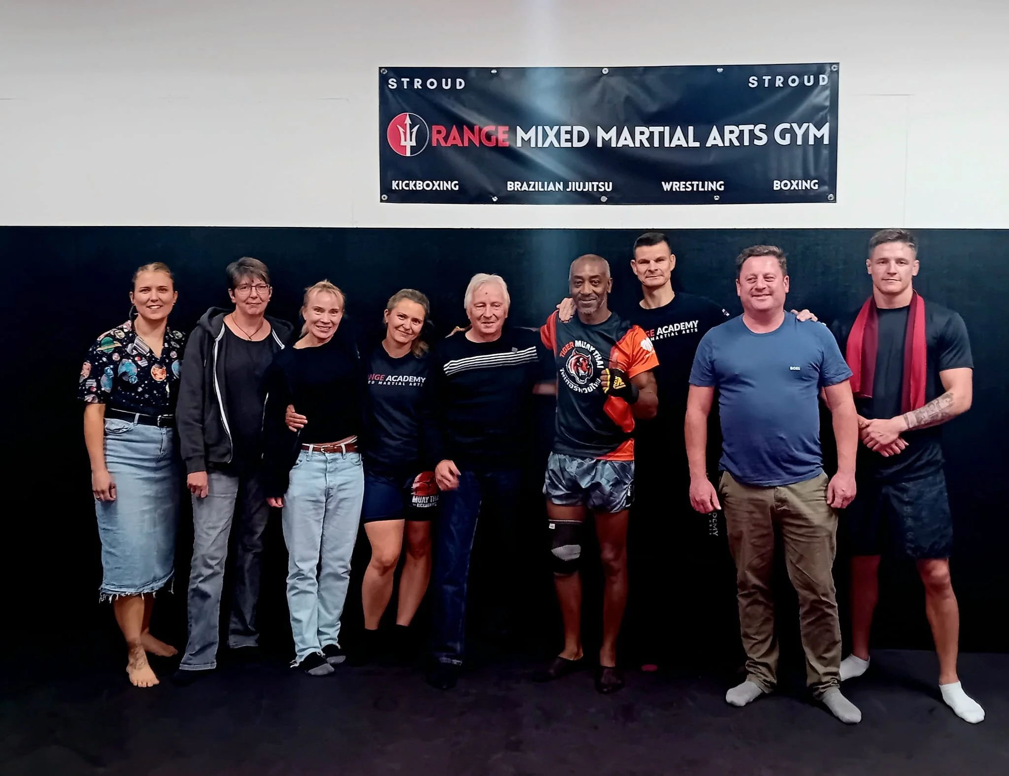 Group of nine people standing together at a martial arts gym, some smiling, with a banner overhead that reads 'Stroud Range Mixed Martial Arts Gym' and lists activities such as kickboxing, Brazilian jiu jitsu, wrestling, and boxing.