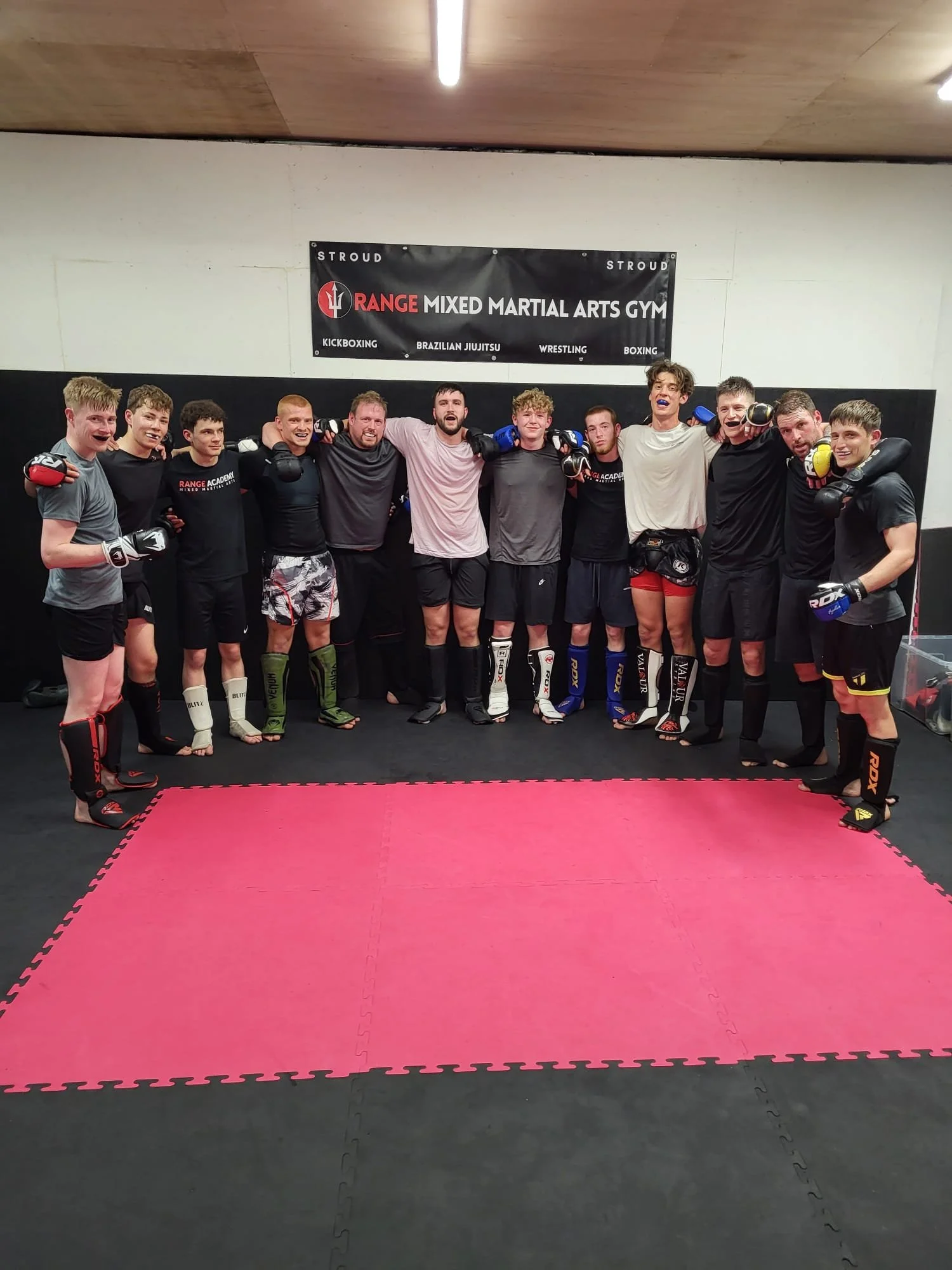 Group of martial arts students and instructor standing in a gym, smiling and posing with their arms around each other. They are wearing martial arts gear, including gloves and shin guards. A banner for 'Range Mixed Martial Arts Gym' is hanging on the