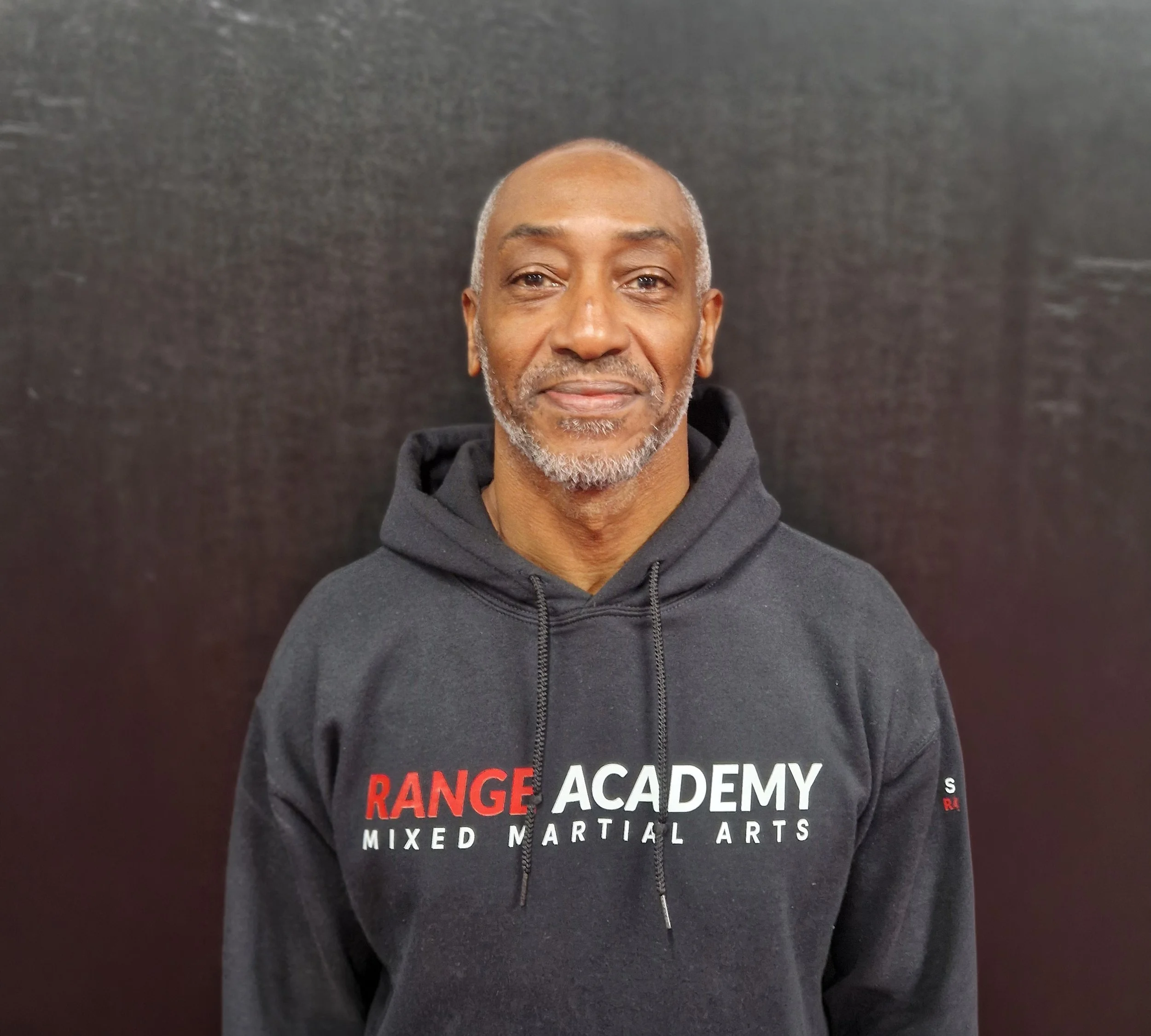 A man with a shaved head and gray beard wearing a black hoodie with the text 'Range Academy' on it, standing against a plain dark background.