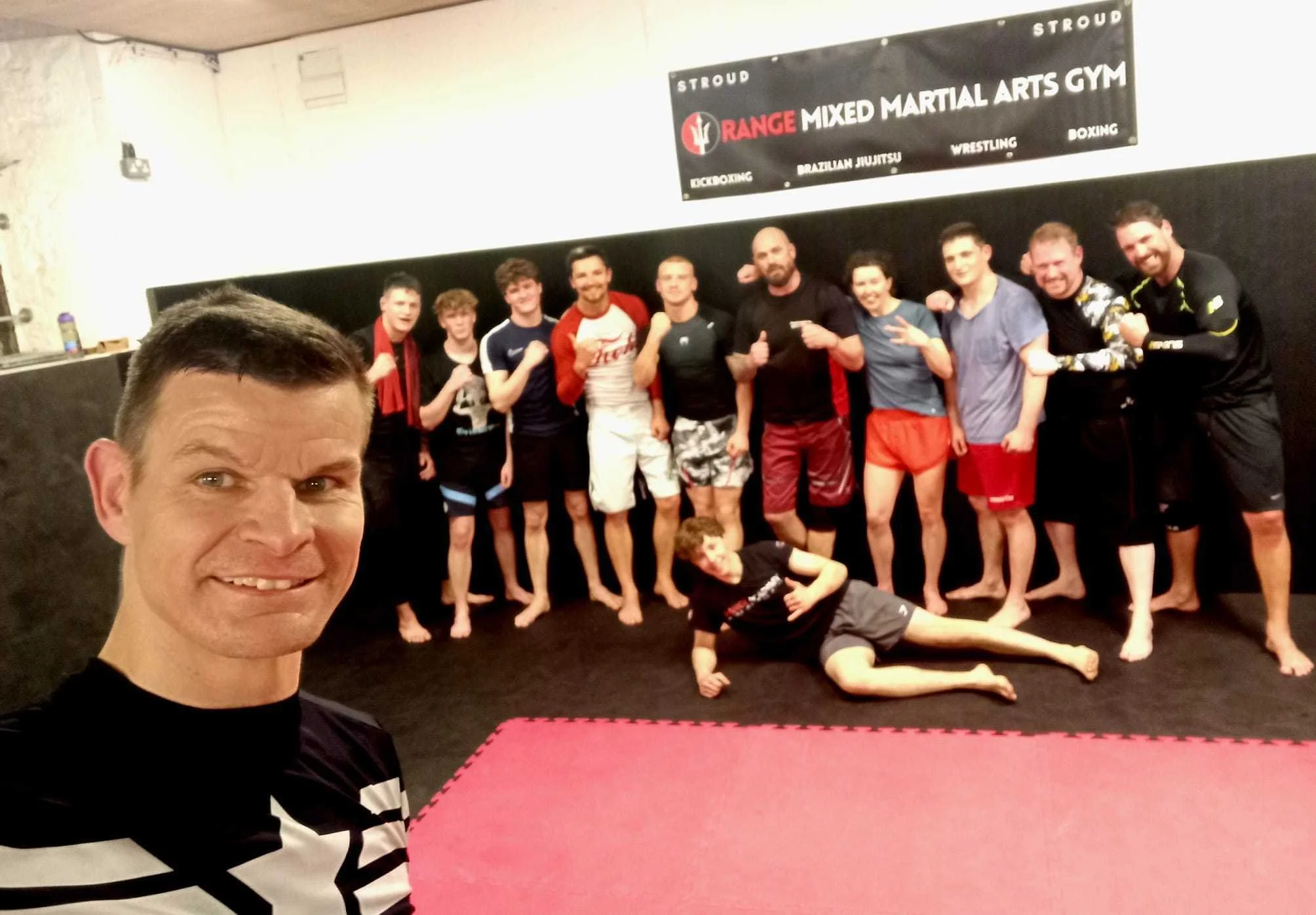 Group of people posing for a photo in a martial arts gym. The background has a sign for Orange Mixed Martial Arts Gym, with categories like Brazilian Jiu Jitsu, Wrestling, Boxing, and Kickboxing. The man in the front is taking a selfie, and others are standing or lying on the floor, some making martial arts gestures or flexing.