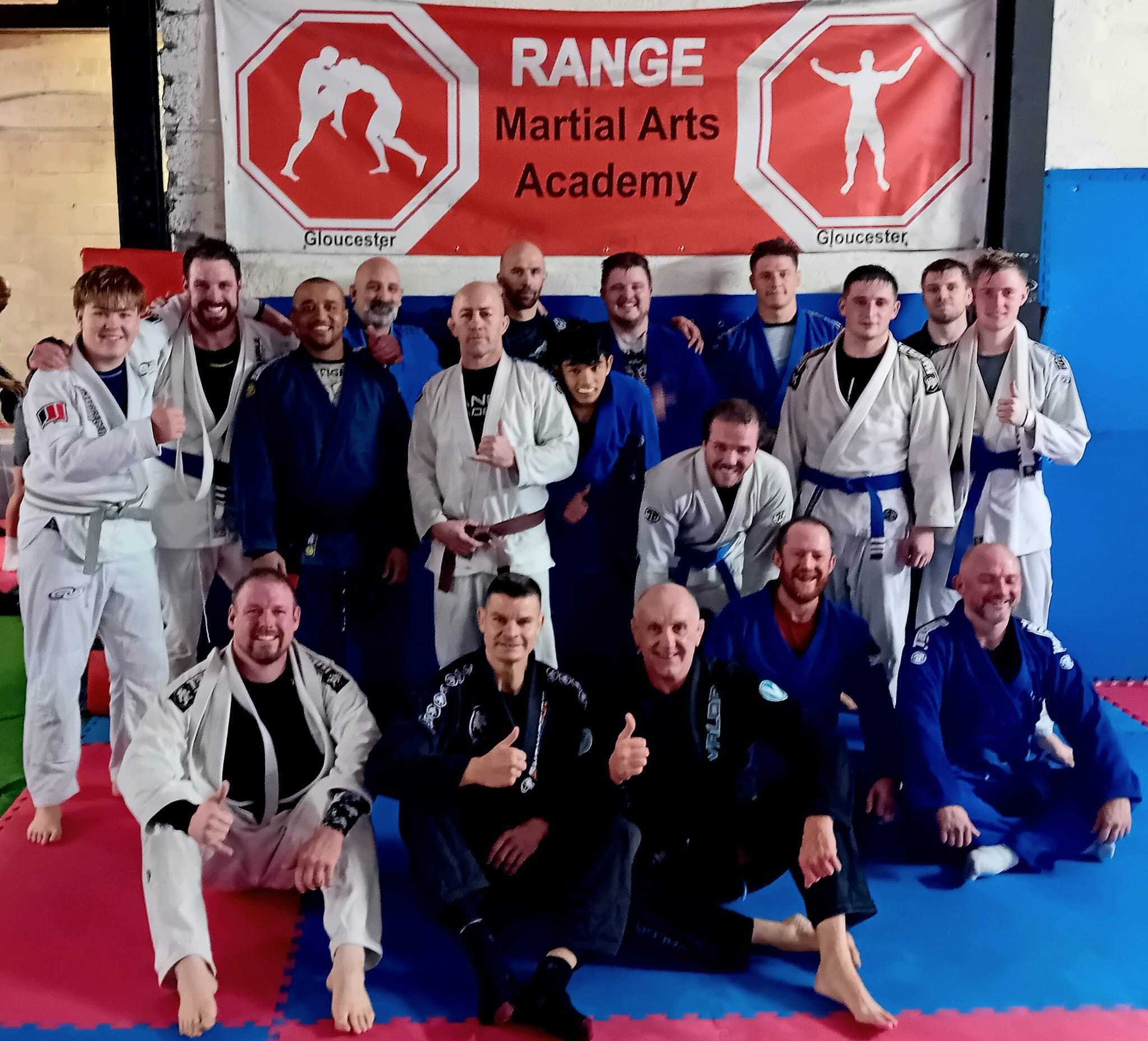Group of martial arts students and instructors in gi uniforms, some giving thumbs up, posing in front of a red and white banner that reads "Range Martial Arts Academy" with images of martial arts silhouettes and the word "Gloucester".