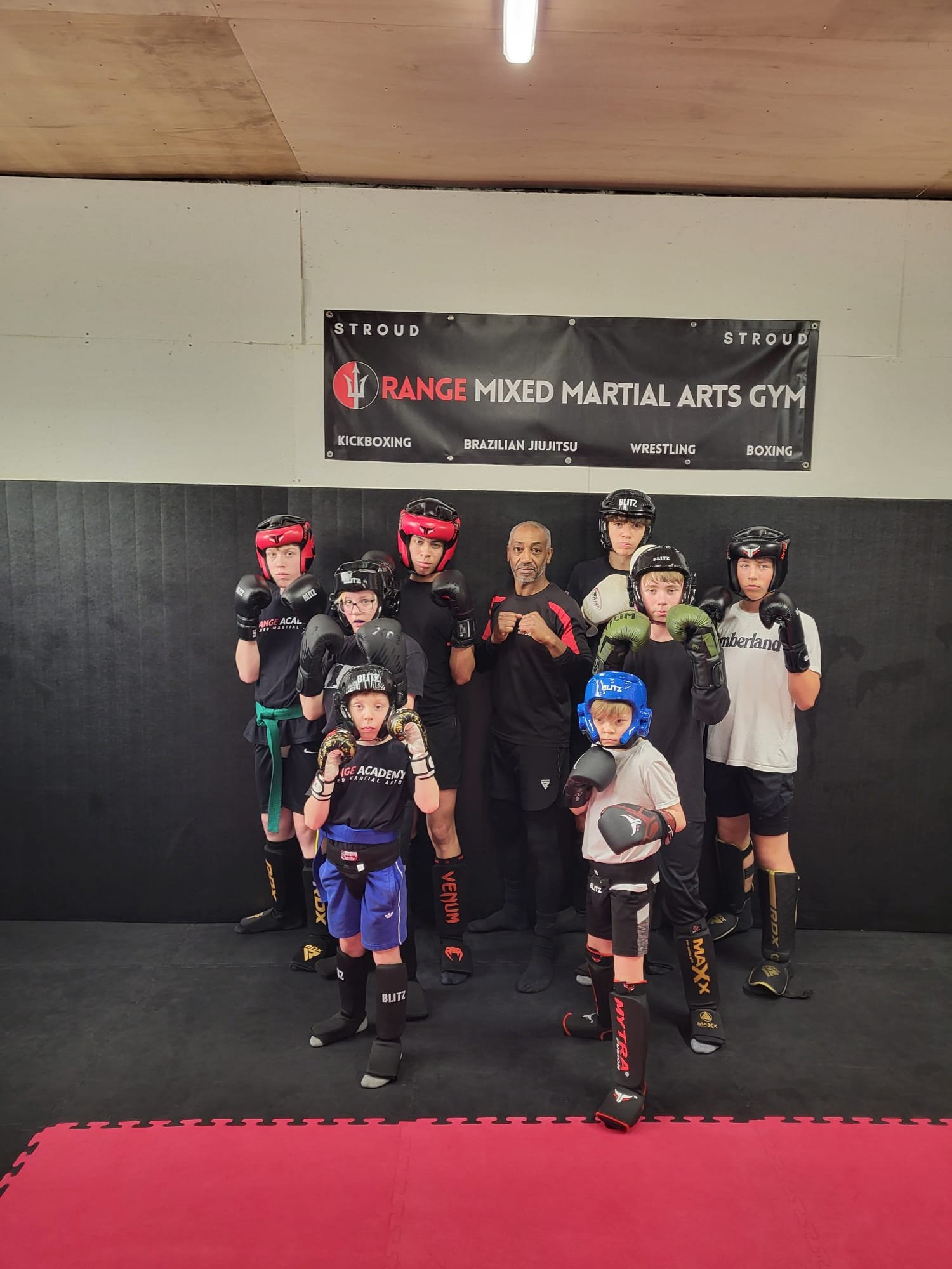 Group of children in martial arts gear with an instructor in a martial arts gym. They are standing in front of a black wall and a banner that reads 'Range Mixed Martial Arts Gym' with disciplines like kickboxing, Brazilian Jiu Jitsu, wrestling, and boxing listed. The children are wearing boxing gloves, helmets, and martial arts uniforms, and some are posing with fists raised.
