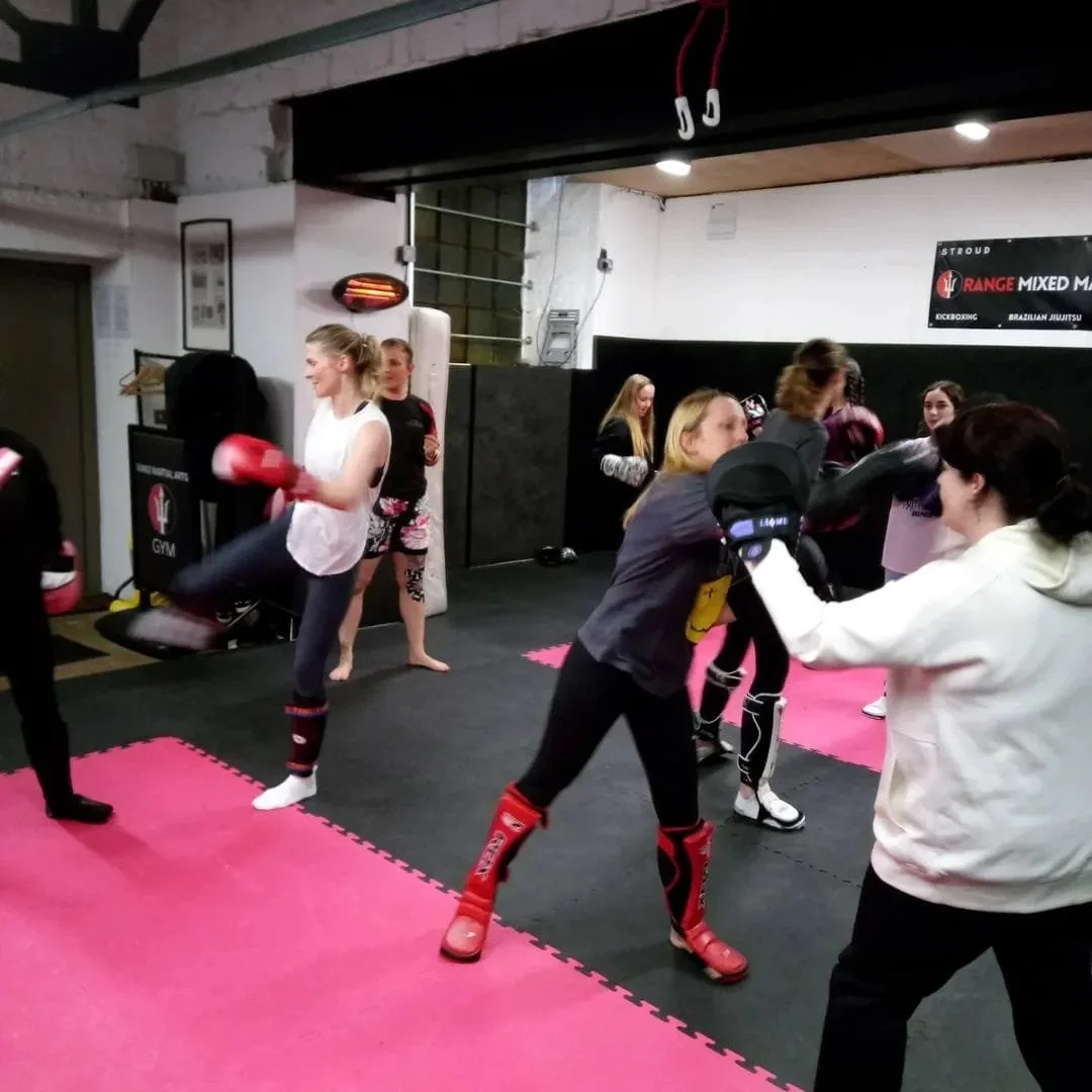 Women practicing kickboxing together in a gym, some wearing protective gear and gloves.