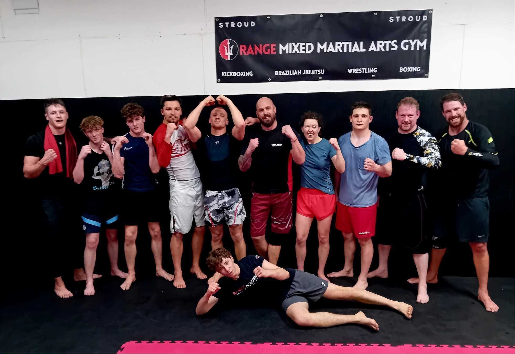 Group of martial arts practitioners posing in a gym, some flexing muscles, with a banner reading 'Range Mixed Martial Arts Gym' in the background.