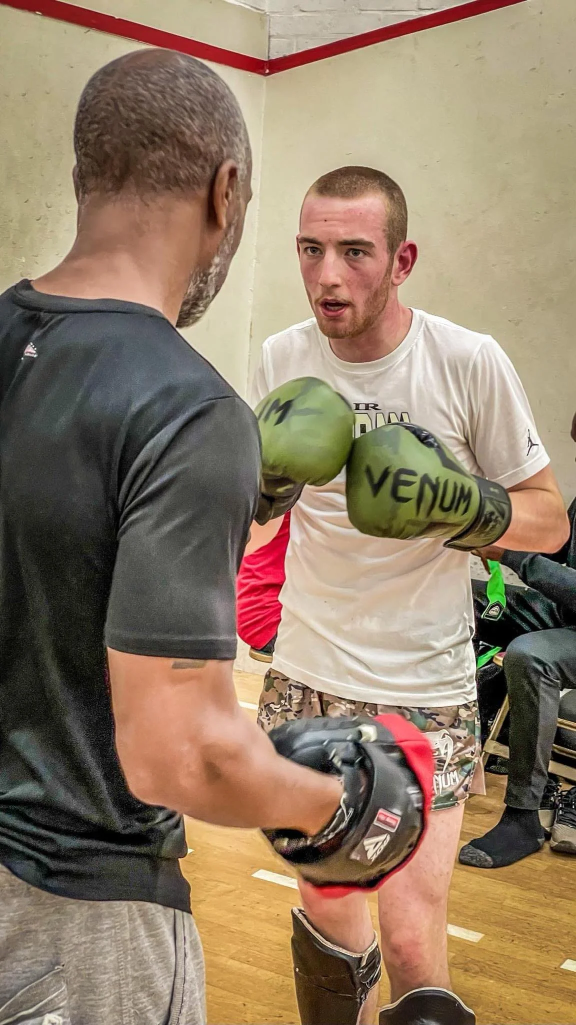 Two men in a boxing gym, one wearing boxing gloves and shorts, the other wearing a black shirt and gray sweatpants, are practicing boxing.