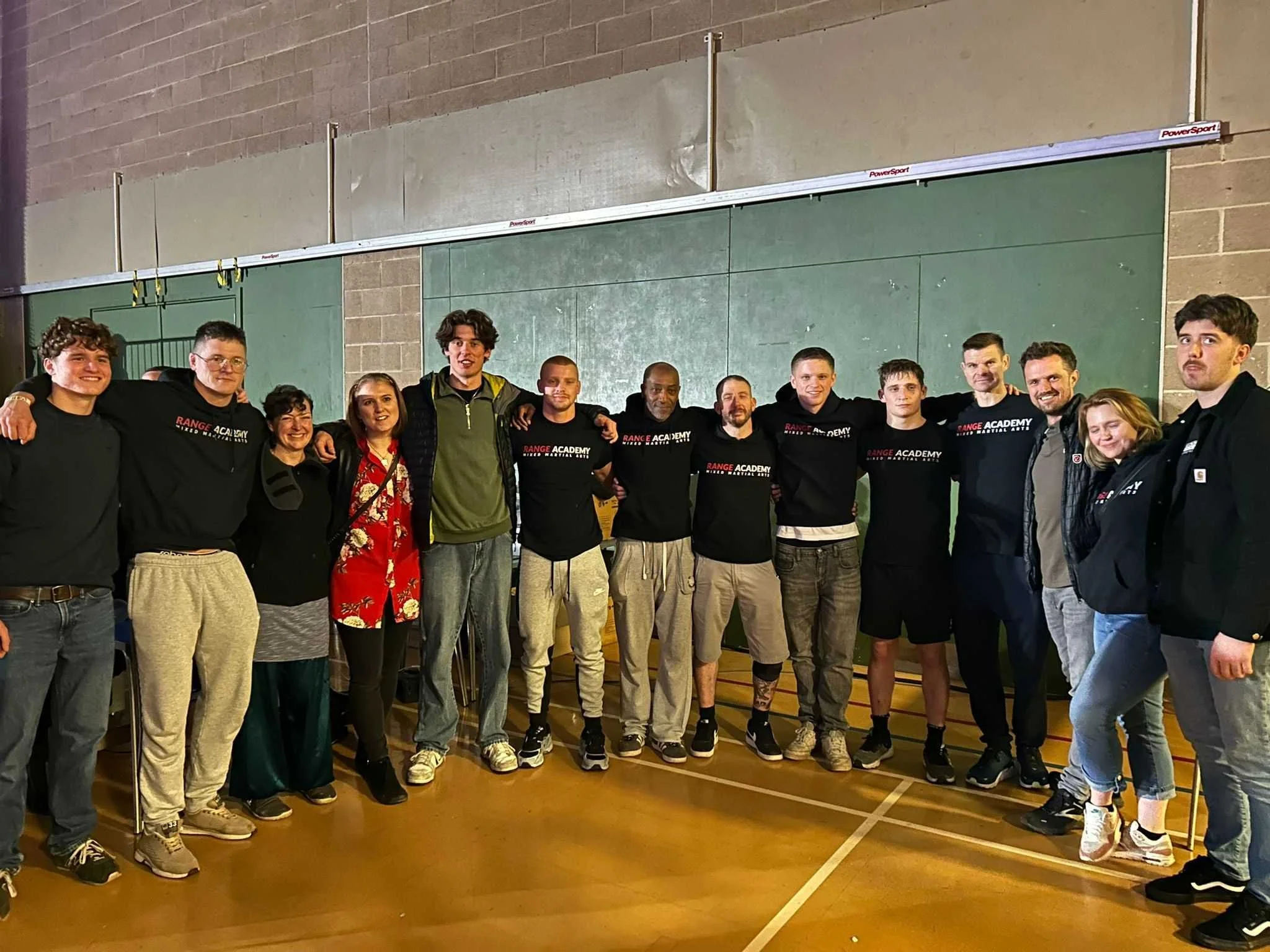 Group of people standing together indoors in front of a green chalkboard and brick wall, some wearing black T-shirts with 'Range Academy' logo.