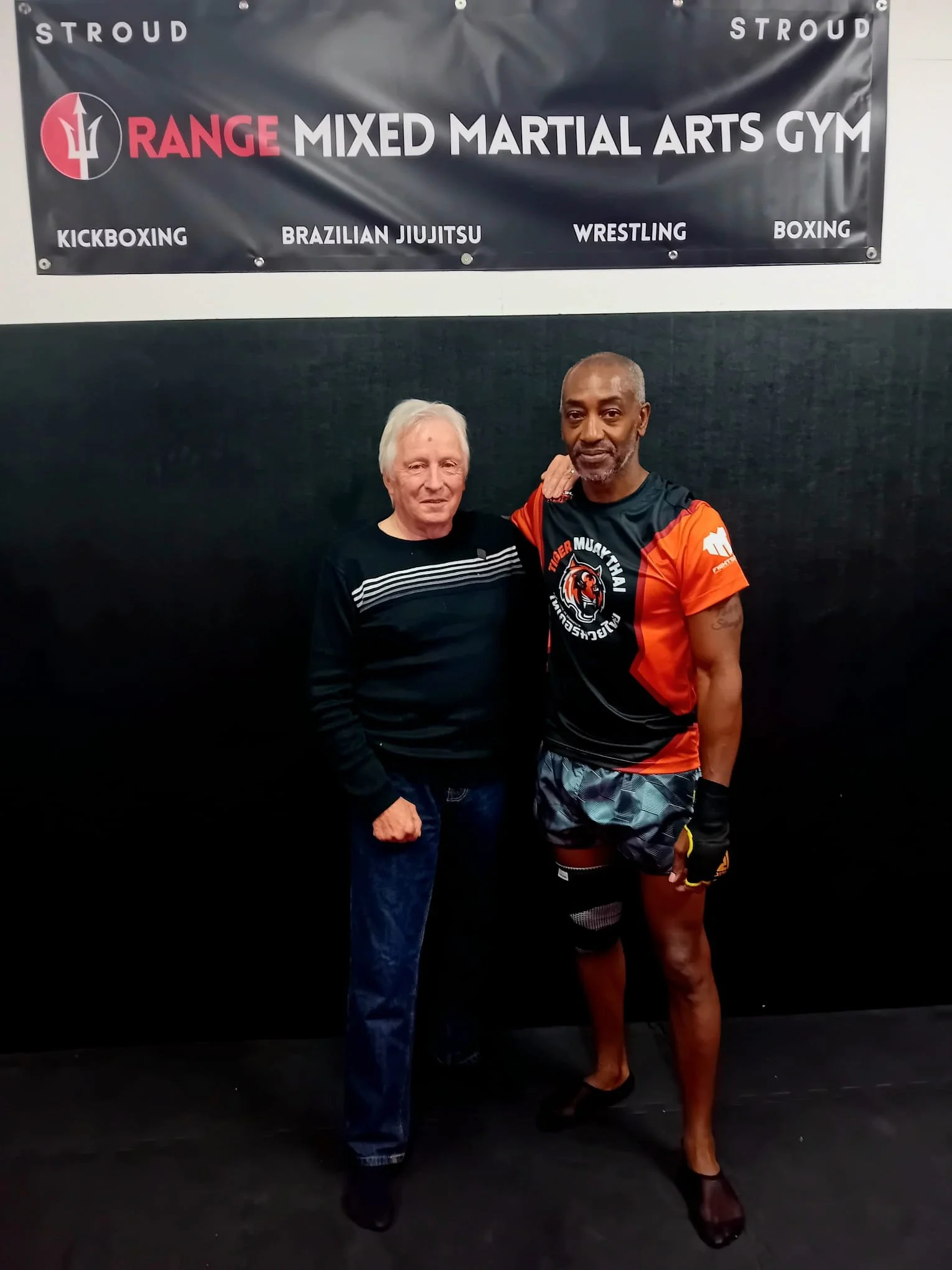 Two men standing side by side in a martial arts gym, one older man and one younger man, posing for a photo. The gym has a black wall with a banner that reads 'Range Mixed Martial Arts Gym' with additional martial arts disciplines listed, including ki
