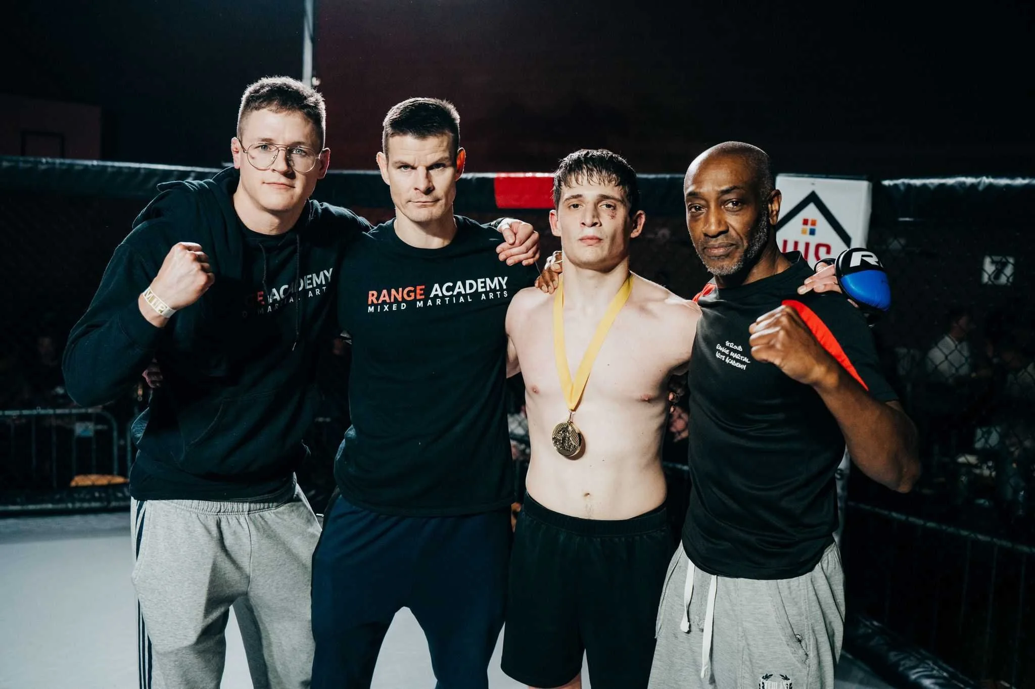 Four male mixed martial arts fighters inside an octagon cage, celebrating after a match, with one shirtless athlete wearing a gold medal.