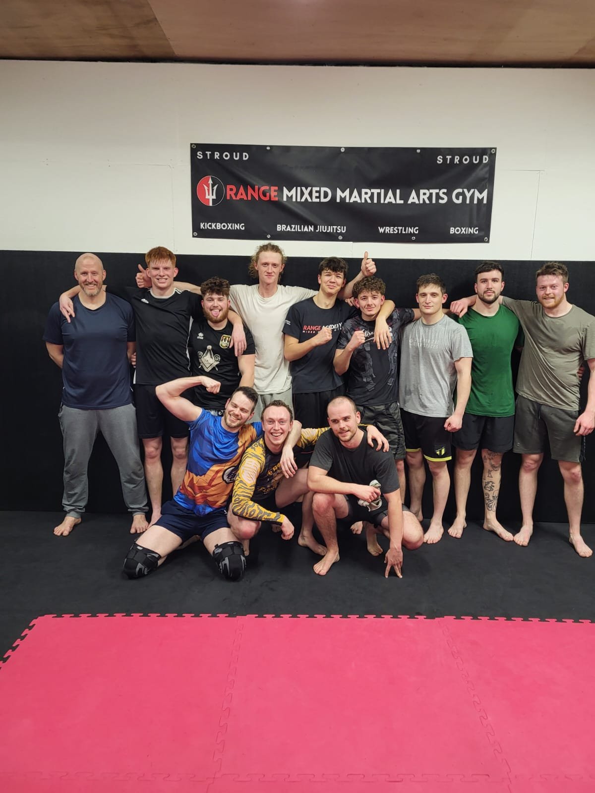 Group of people posing together in a martial arts gym, some flexing muscles and smiling, with a black banner that reads 'Range Mixed Martial Arts Gym' and lists various martial arts disciplines.