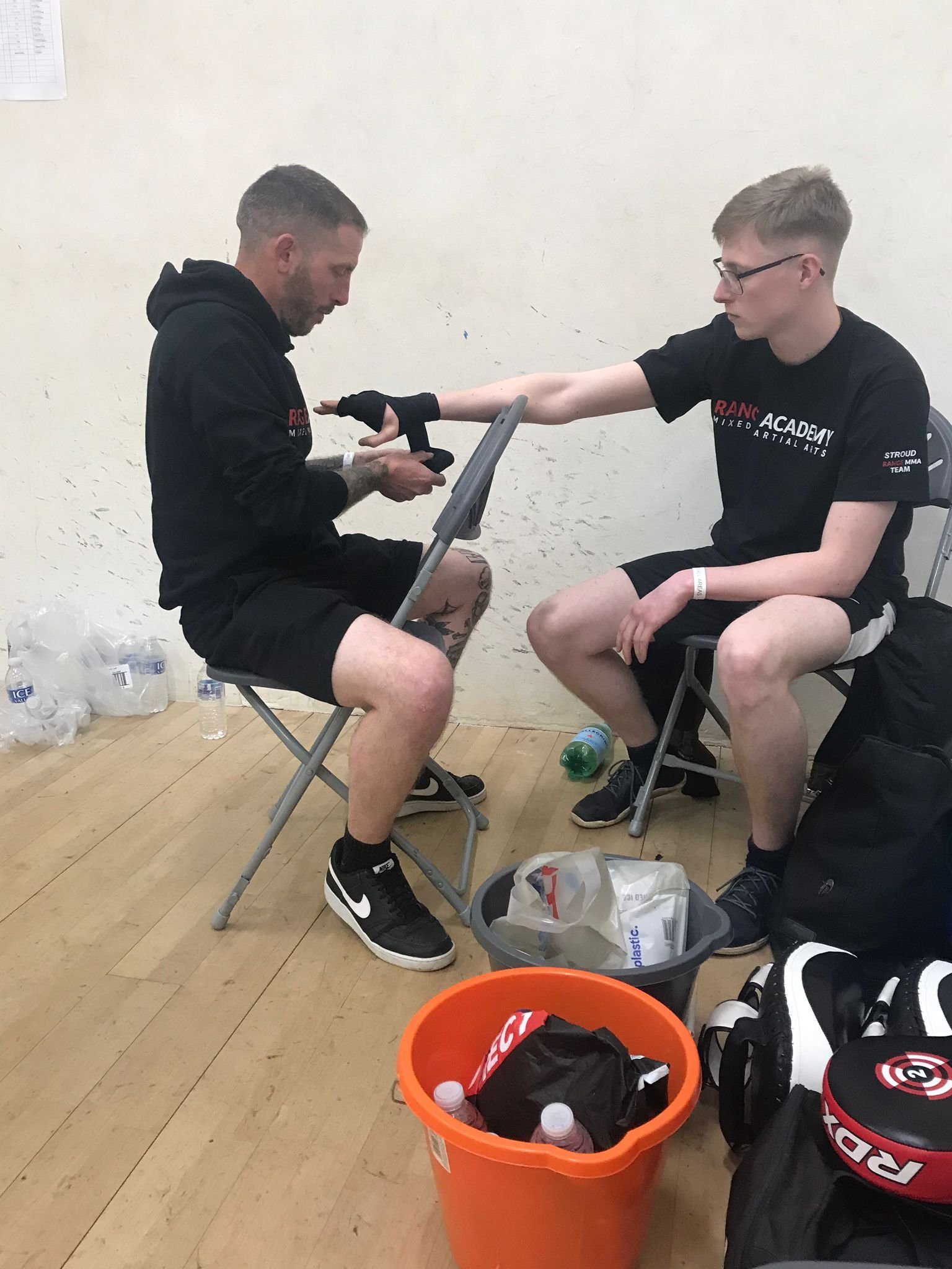 Two young men sitting on chairs against a plain wall. One man is looking at his phone, and the other is reaching out with his arm. They are surrounded by water bottles, bags, and sports equipment in the floor and in a large orange container.