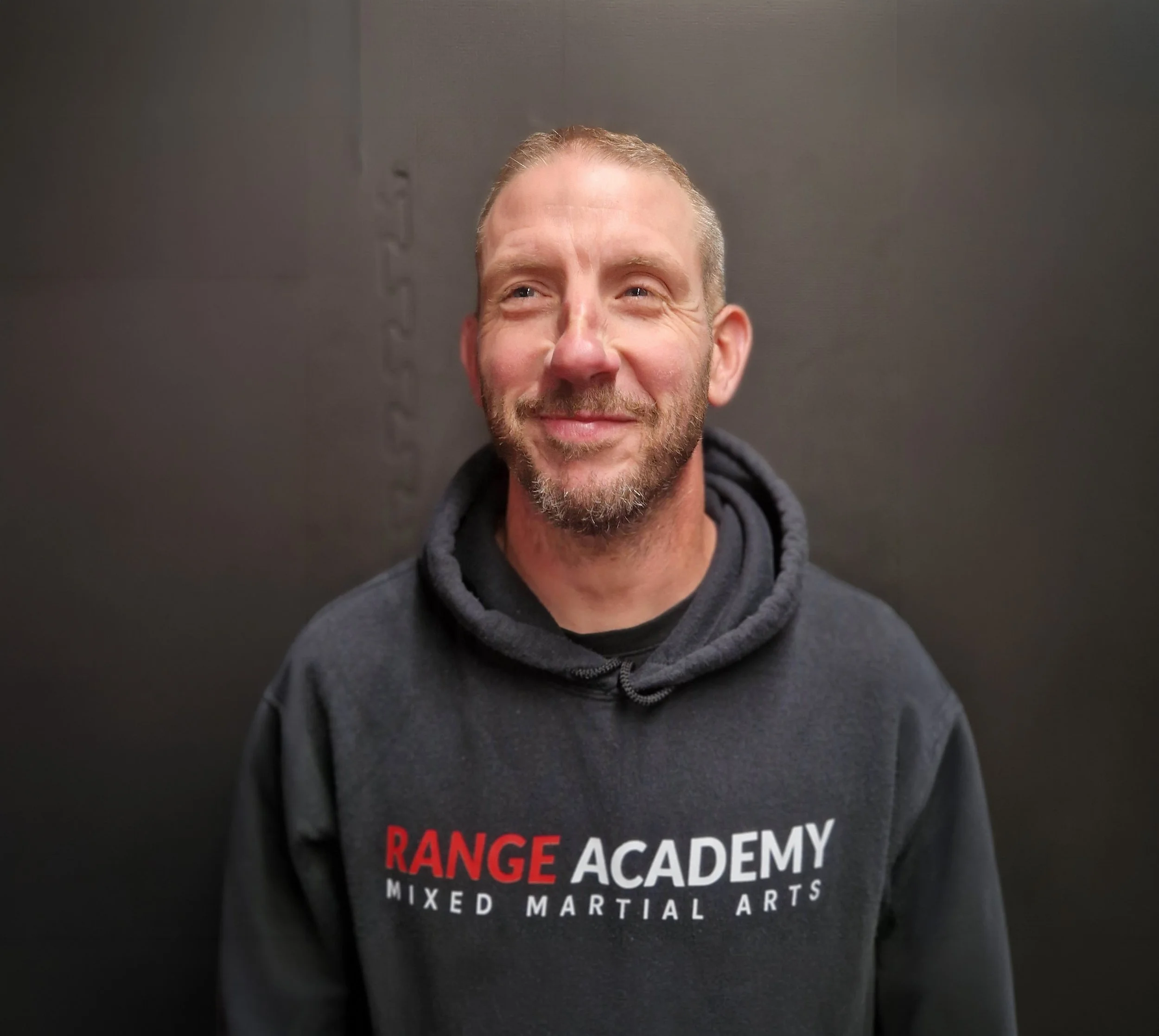 Close-up of a smiling man with a beard, wearing a dark hoodie, standing in front of a black wall. Part of a fitness or martial arts class logo is visible on his shirt.