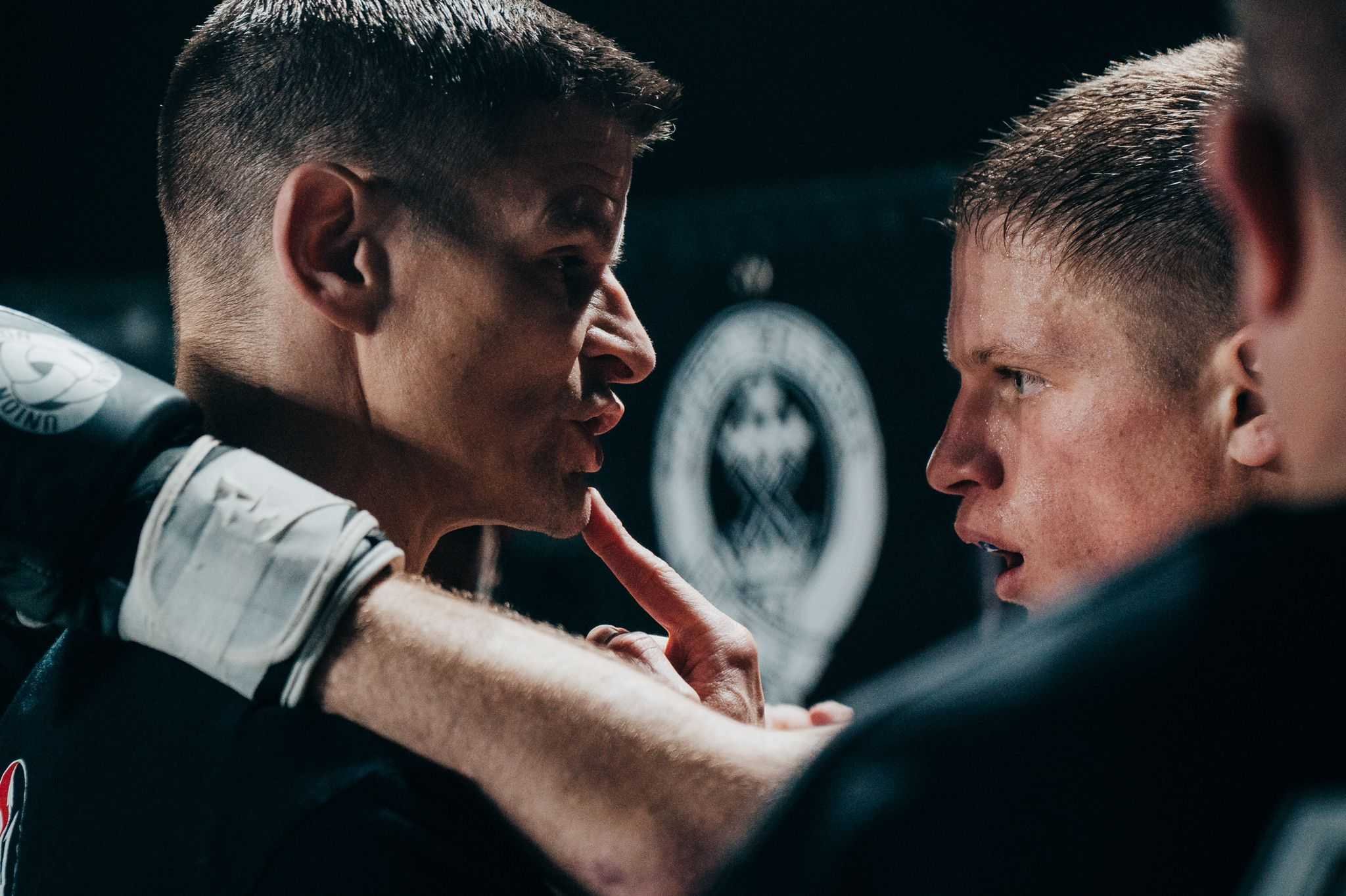 Two men in a close confrontation, one with a finger on the other's lips, in a serious discussion, with a blurred logo in the background.