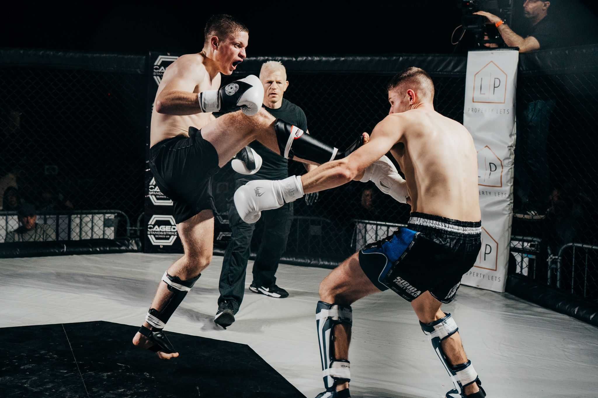 Two mixed martial artists engaging in a fight inside a cage, one delivering a high kick to the other's head while the other blocks with his arms; referee and cameraman observing.