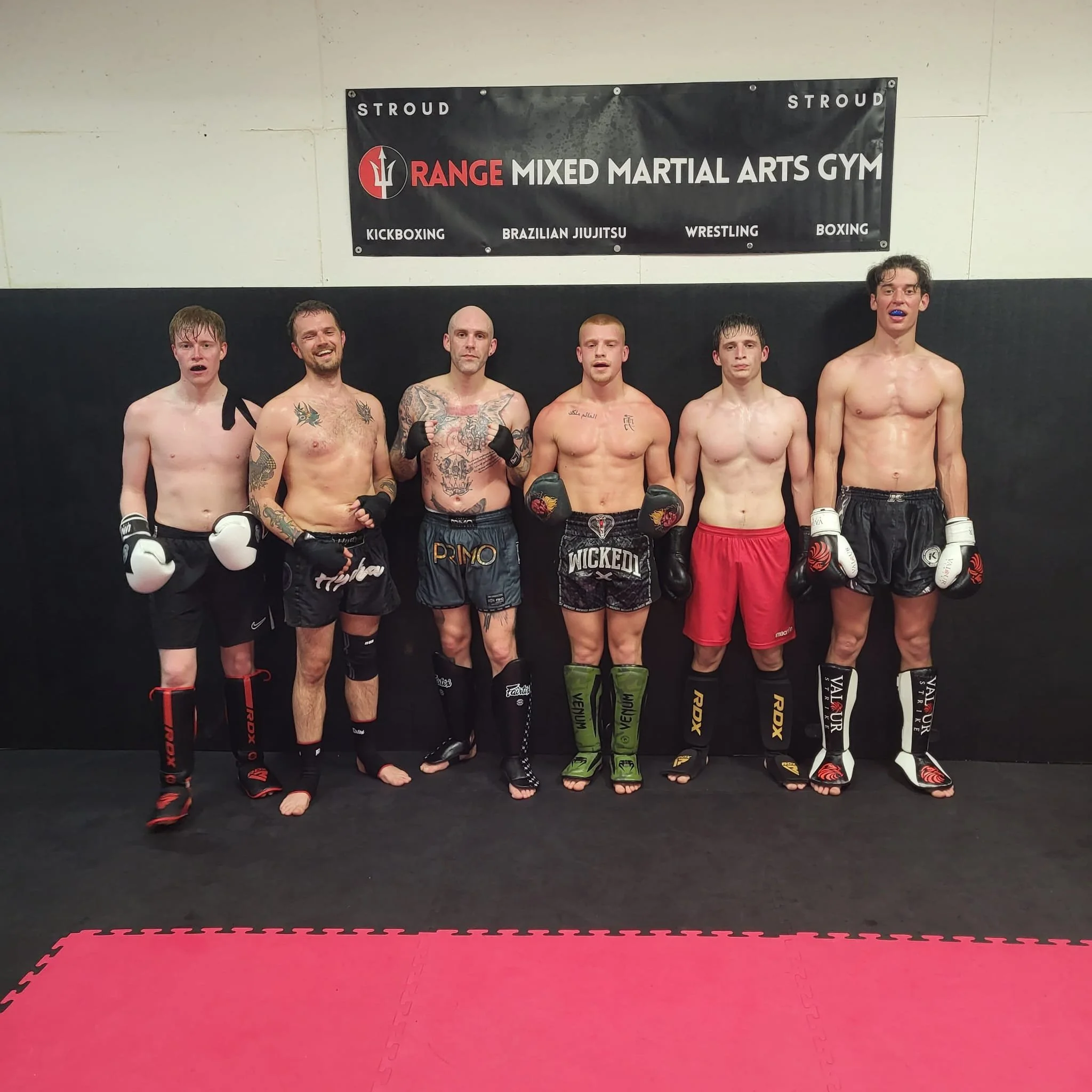 Group of six shirtless men with boxing gloves standing in a martial arts gym, with a black and red banner behind them that reads 'Range Mixed Martial Arts Gym' and lists activities like kickboxing, Brazilian jiu-jitsu, wrestling, and boxing. The men 