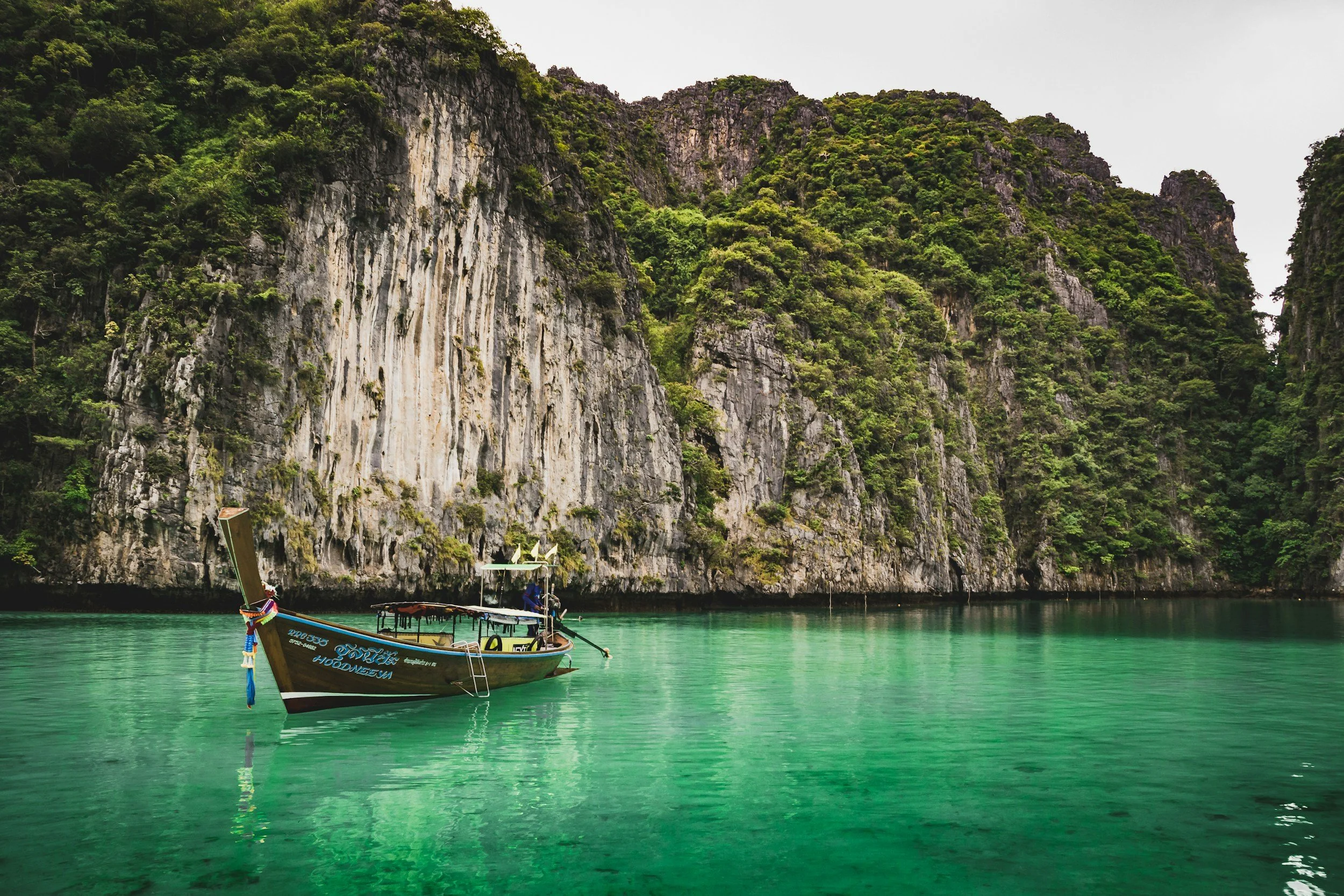 Bateau traditionnel en bois flottant sur une eau turquoise avec des falaises verdoyantes en arrière-plan.