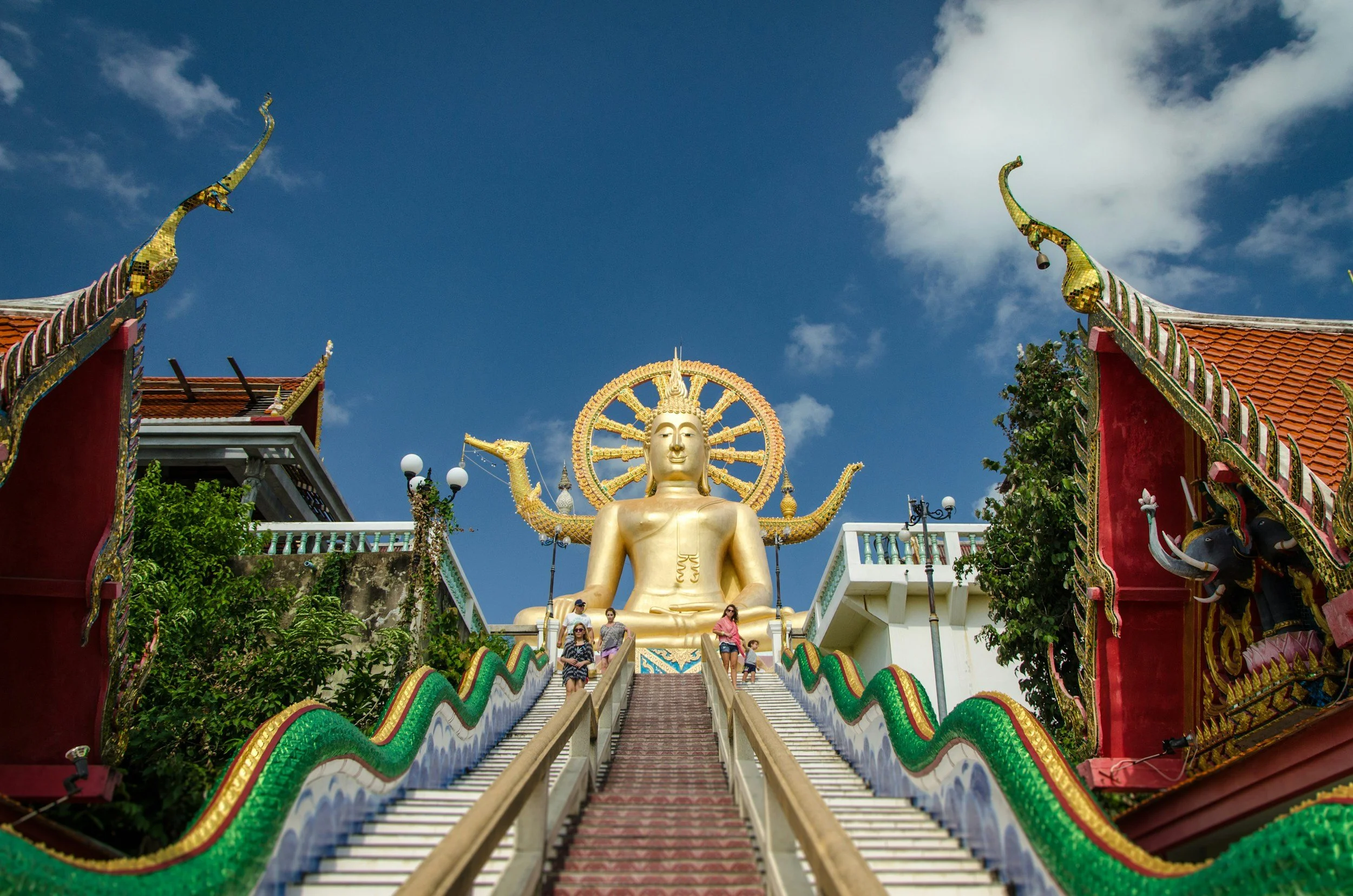 Escalier menant à une grande statue dorée de Bouddha, avec un halo et des serpents décoratifs sur les côtés, dans un temple thaïlandais.