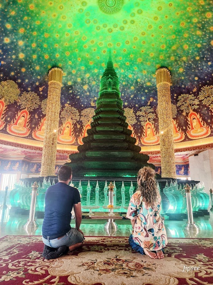 Deux personnes, un homme et une femme, sont agenouillées devant un grand stupa dans une pièce richement décorée avec un plafond coloré de motifs lumineux et de Bouddhas dorés entourés de végétation. Ils prient ou méditent dans un lieu religieux ou spirituel.