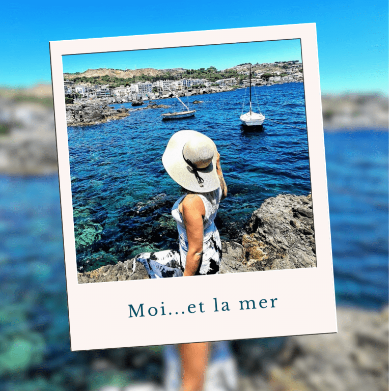 Une femme portant un chapeau blanc assise sur des rochers près de la mer, avec des bateaux à voile et une ville en arrière-plan, ensoleillé.