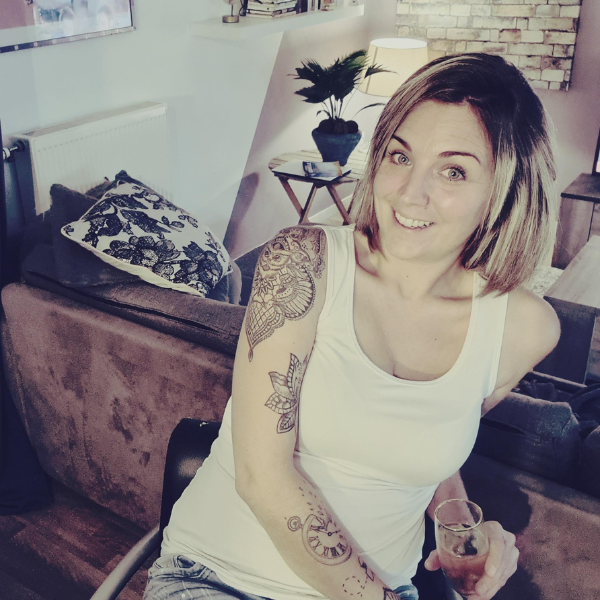 Femme souriante avec tatouages, tenant un verre, à l'intérieur d'une pièce moderne avec un canapé et une plante.