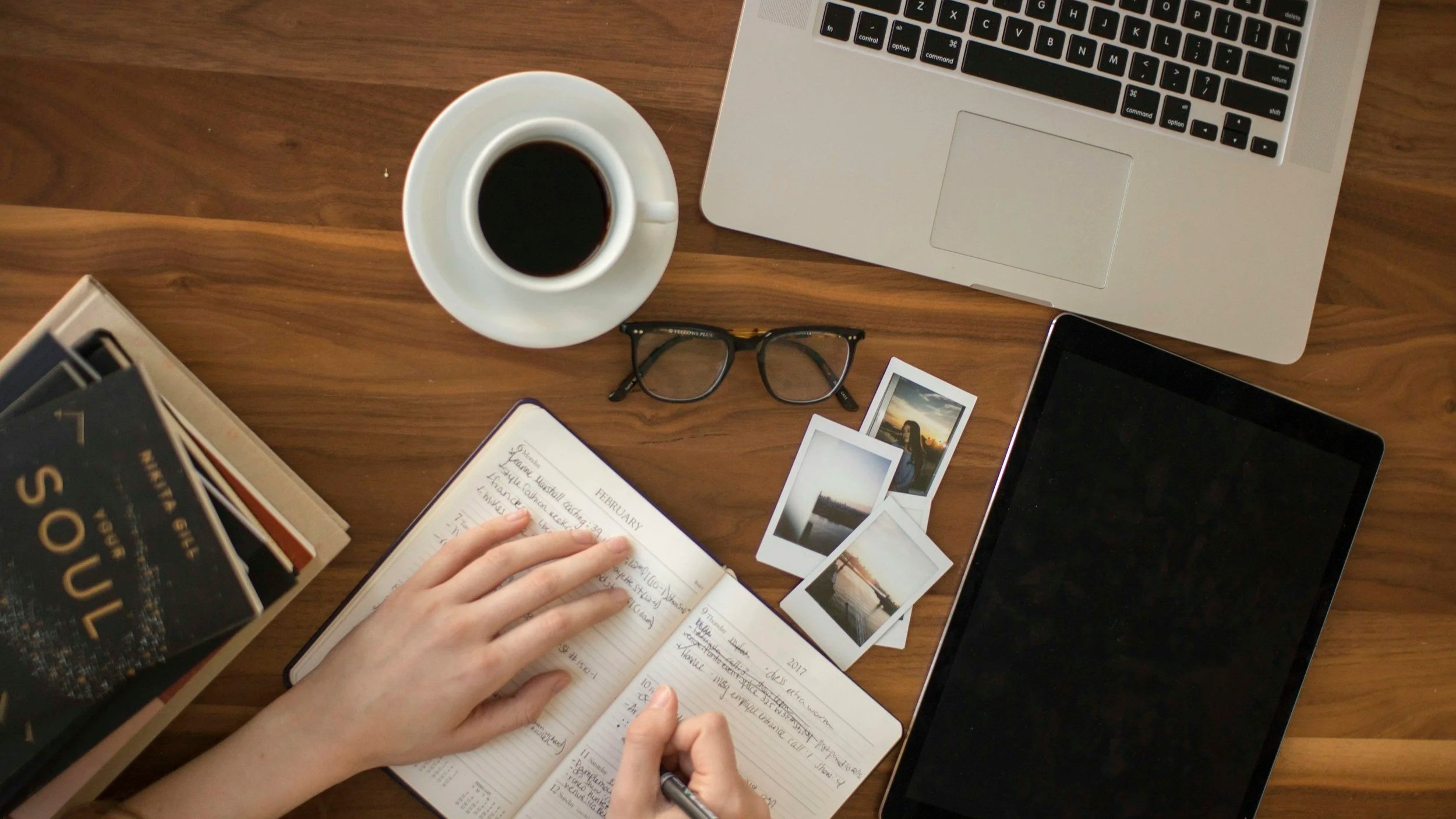 Un bureau en bois avec un ordinateur portable, une tablette, des lunettes, un carnet ouvert, un stylo, un mug de café, des photos instantanées, et une pile de livres.