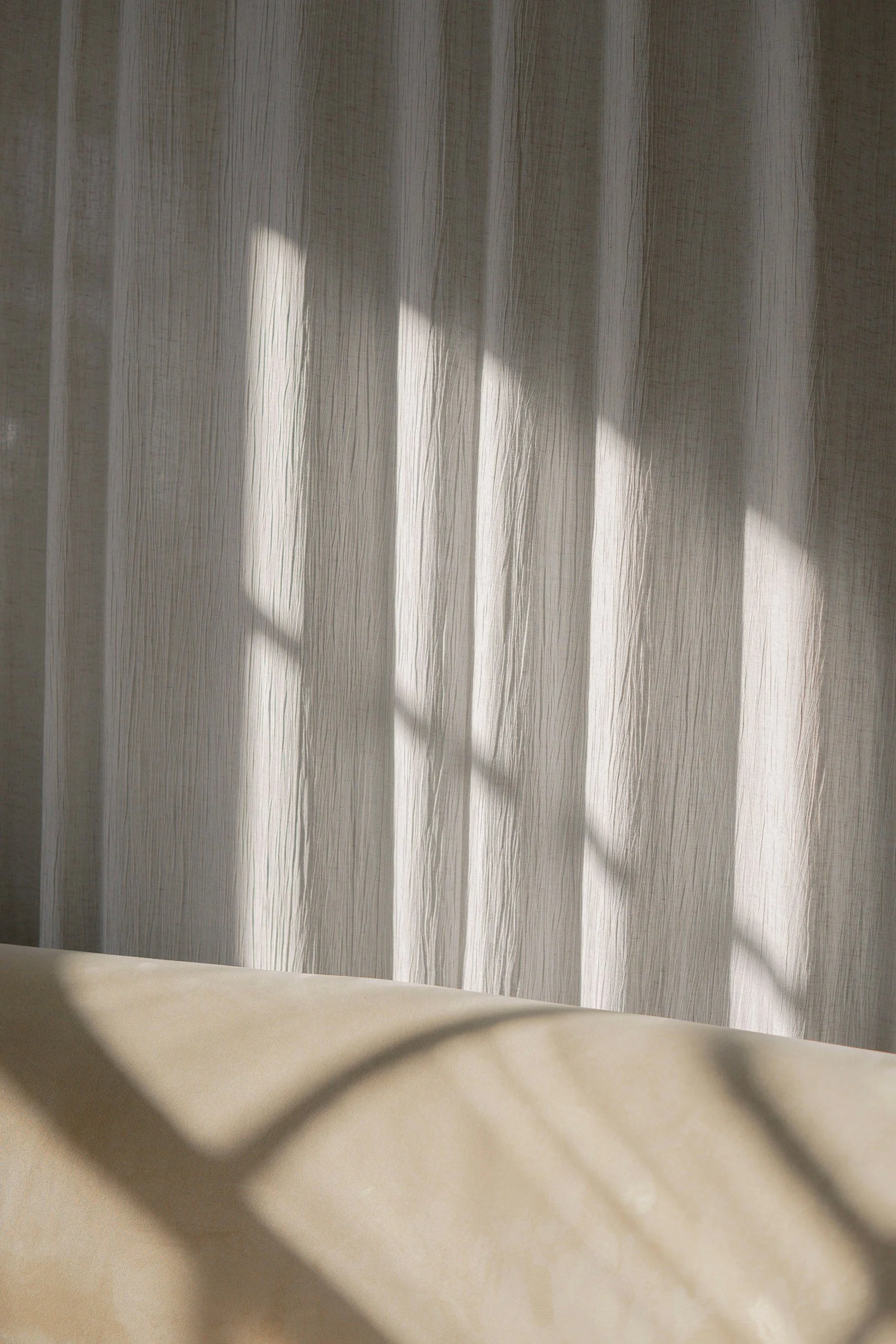 A sheer white curtain with vertical stripes, showing a faint silhouette of a person or object behind it, with sunlight casting shadows through the fabric.